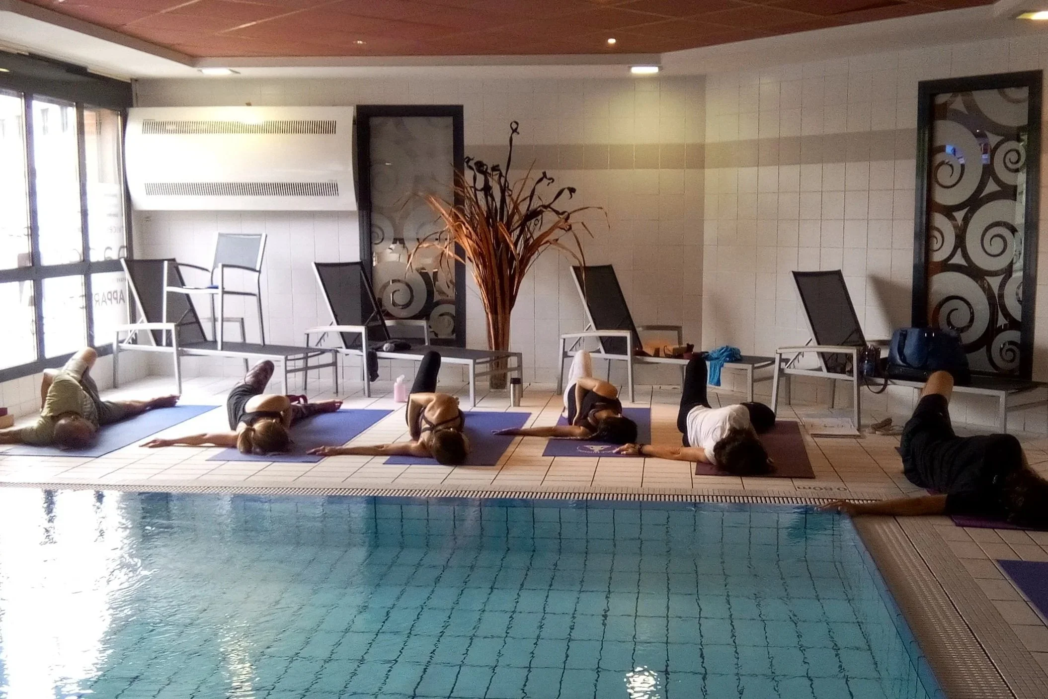Groupe de personnes faisant du yoga près d'une piscine intérieure, allongées sur des tapis de yoga, en position détendue.