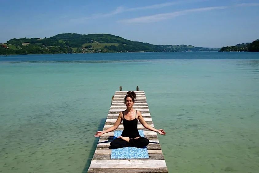Ludivine pratique la méditation en position du lotus sur un tapis en bois au bord d'un lac calme avec une montagne en arrière-plan à Charavines.