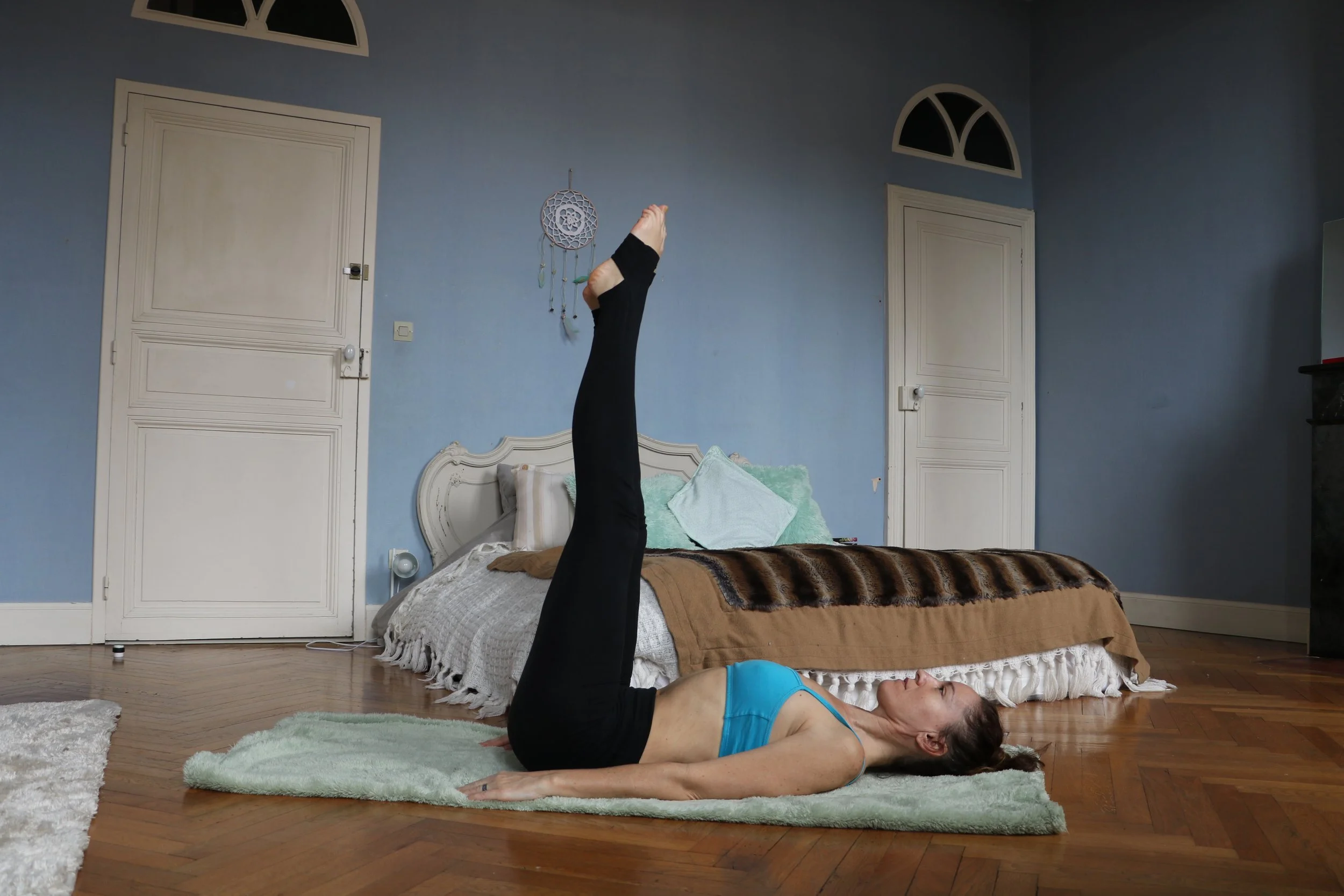 Femme faisant une posture de yoga allongée sur un tapis dans une chambre avec lit et murs bleus. Posture pour mieux dormir.
