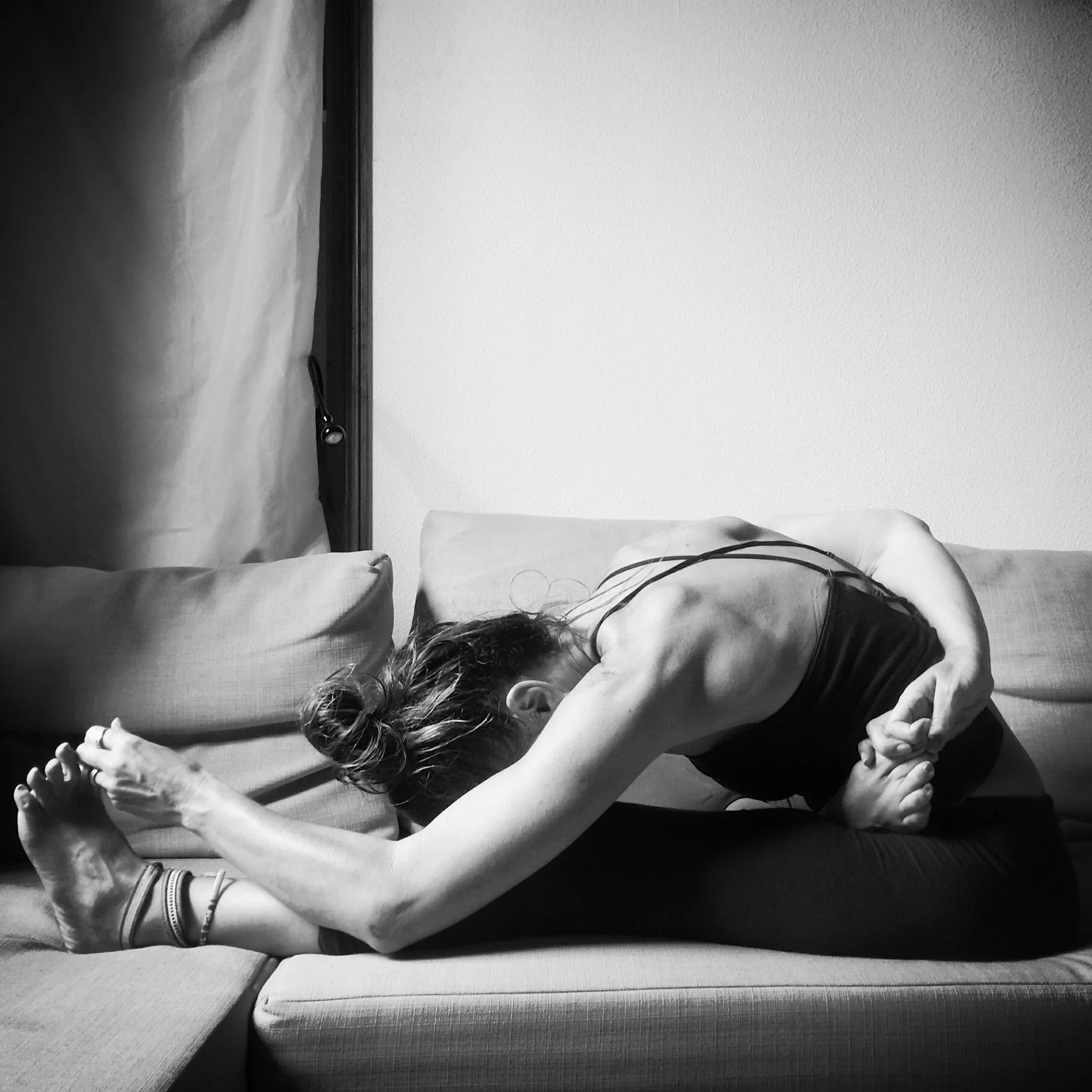 Femme pratiquant le yoga en position de flexion avant, assise sur un canapé, en noir et blanc.