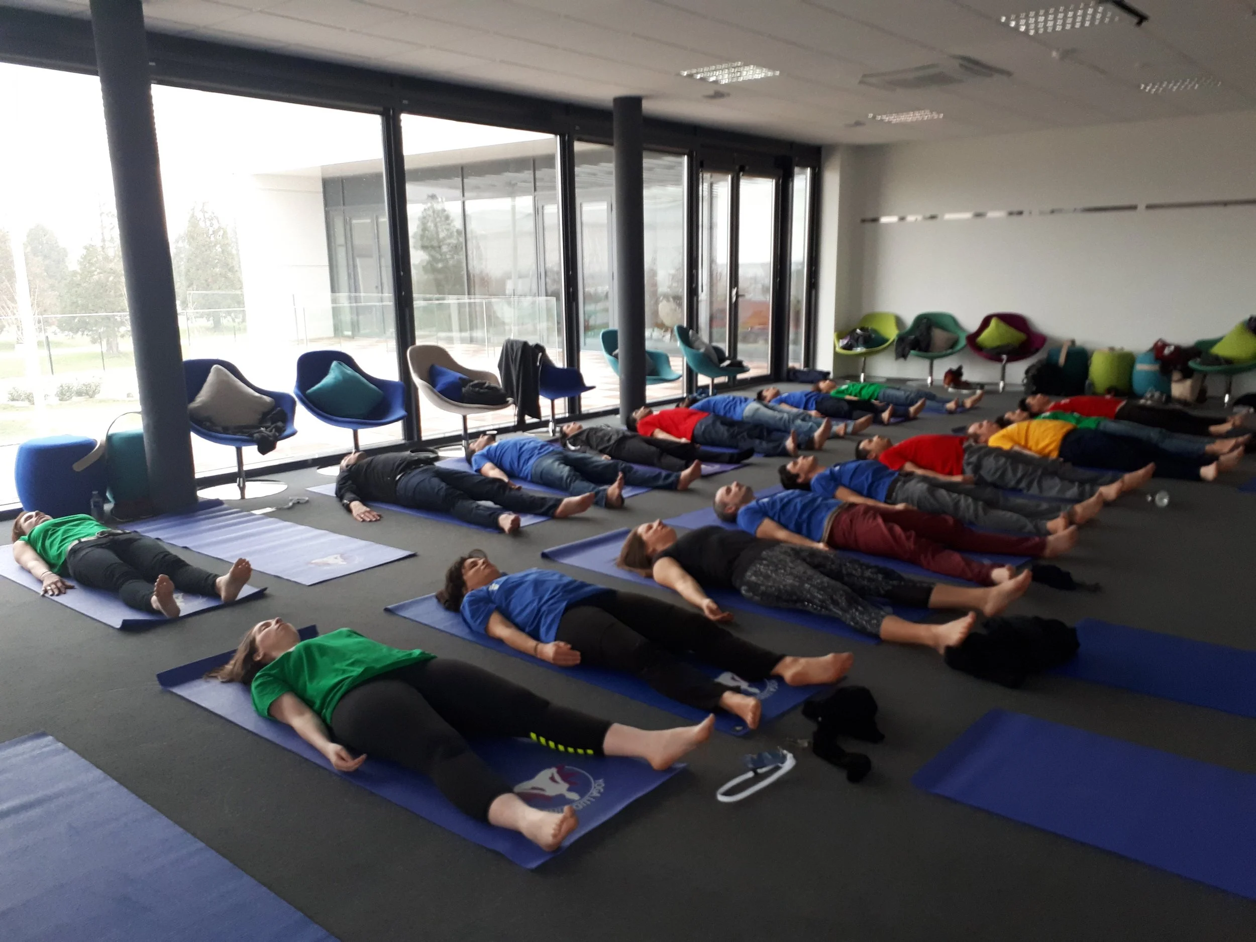 Groupe de personnes participant à une séance de yoga ou de méditation dans une salle intérieure avec des tapis de yoga et des fauteuils près de grandes fenêtres. Ils sont allongés en position de relaxation.