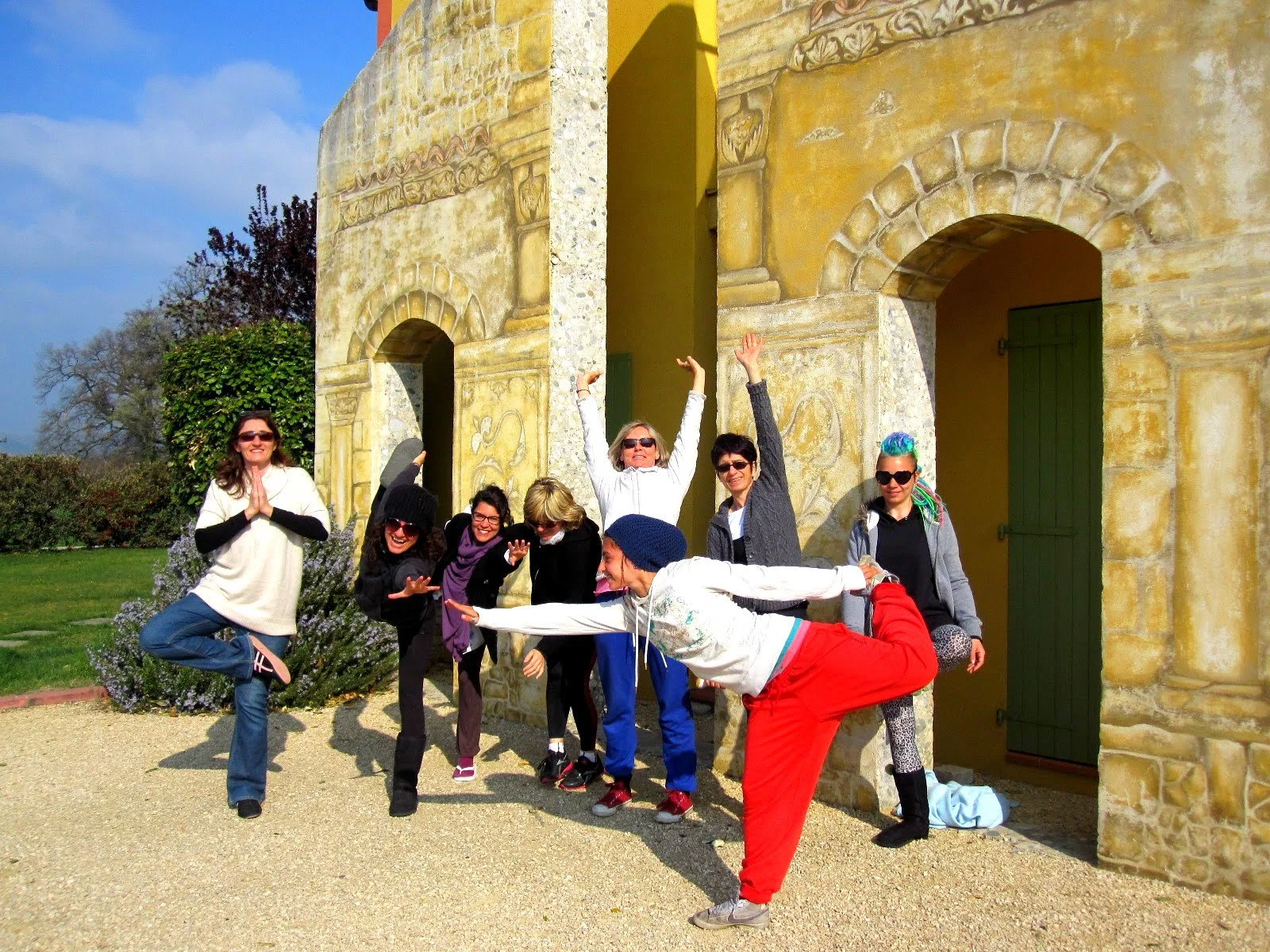 Groupe de huit personnes posant de manière joyeuse devant une vieille façade en pierre avec des arches, en plein jour en plein air, certains faisant semblant de faire du yoga ou de la danse.