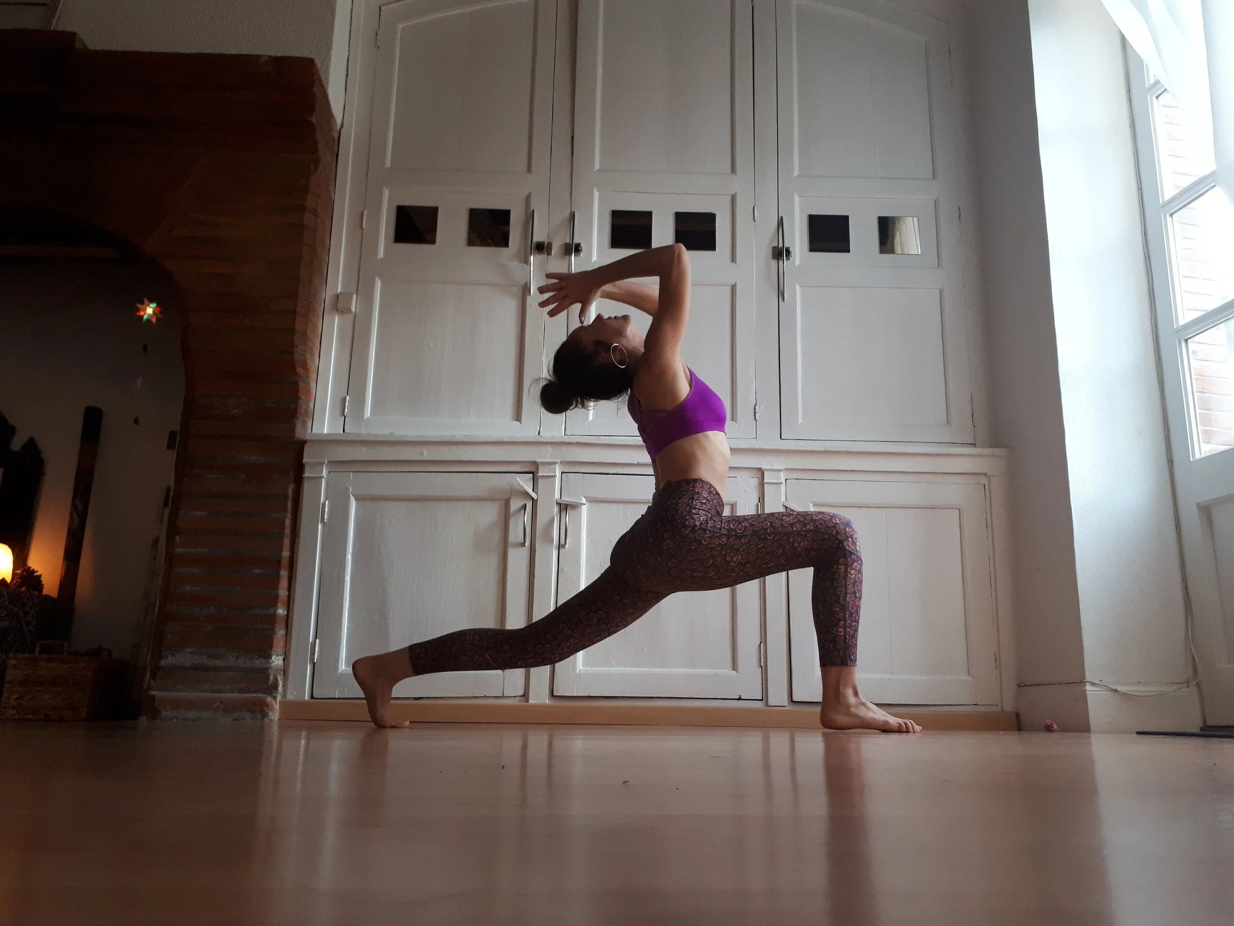 Une femme pratique le yoga dans une pièce lumineuse, faisant une posture en fente avec les mains levées au-dessus de la tête.