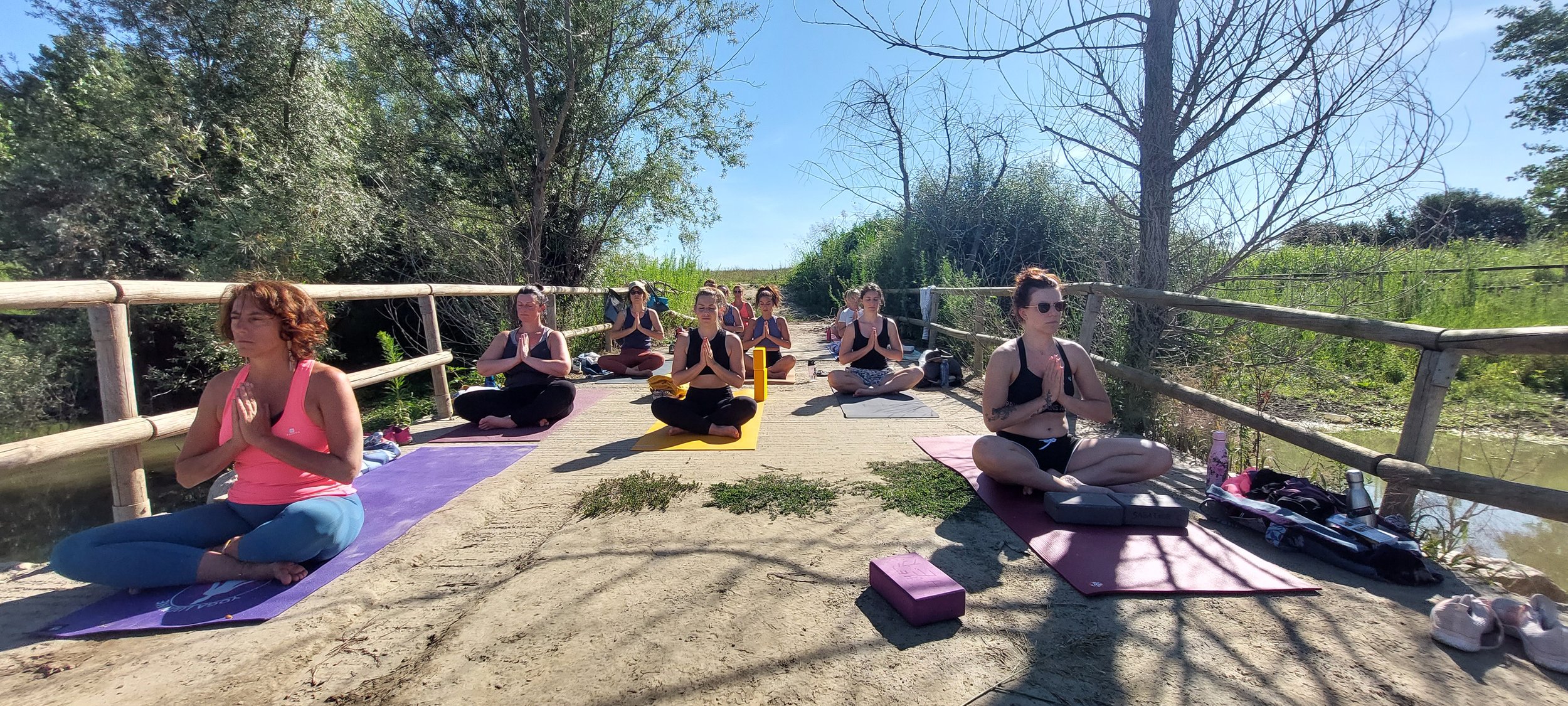 Groupe de personnes pratiquant la méditation en plein air sur un pont en bois, entouré de nature et sous un ciel clair.