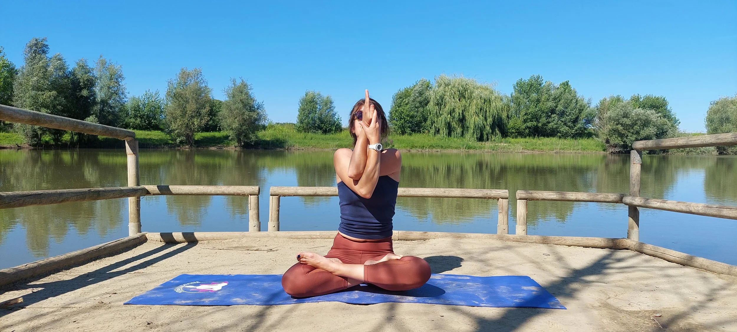 Une femme pratique le yoga en position de lotus sur un tapis bleu, près d'un lac avec des arbres en arrière-plan, par une journée ensoleillée.