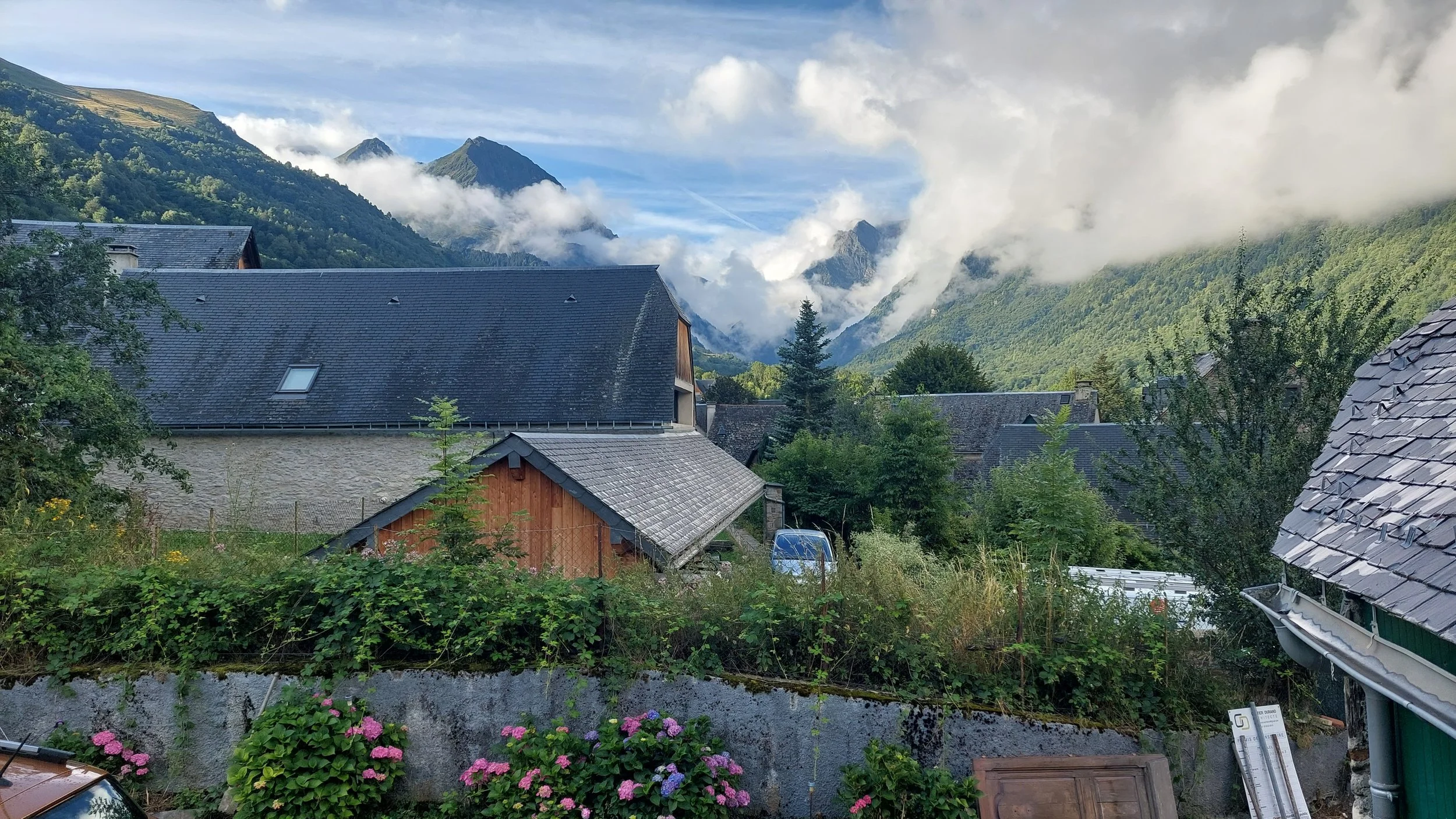 Vue d'un village montagnard avec des maisons en pierre et en bois, entouré de verdure, sous un ciel nuageux avec des montagnes en arrière-plan.