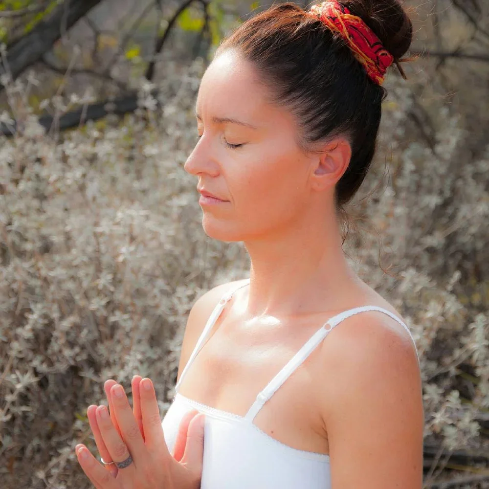 Femme méditant avec les mains jointes, yeux fermés, dans un lieu extérieur avec des arbustes flous en arrière-plan.