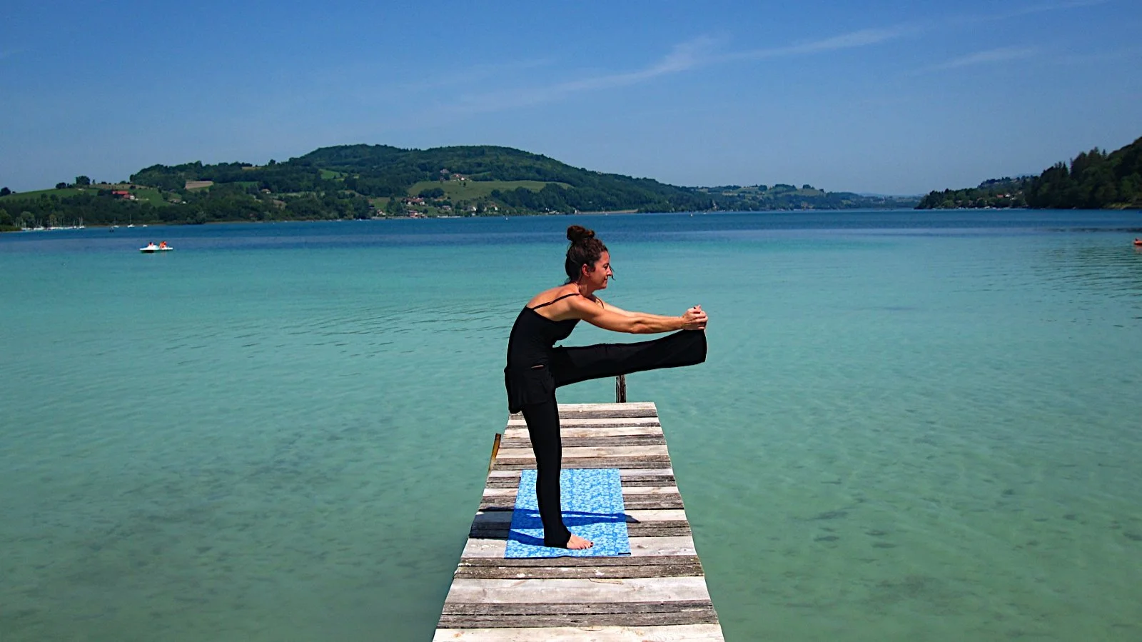 Femme pratiquant le yoga sur une jetée en bois au bord d'un lac calme entouré de collines verdoyantes, sous un ciel clair.