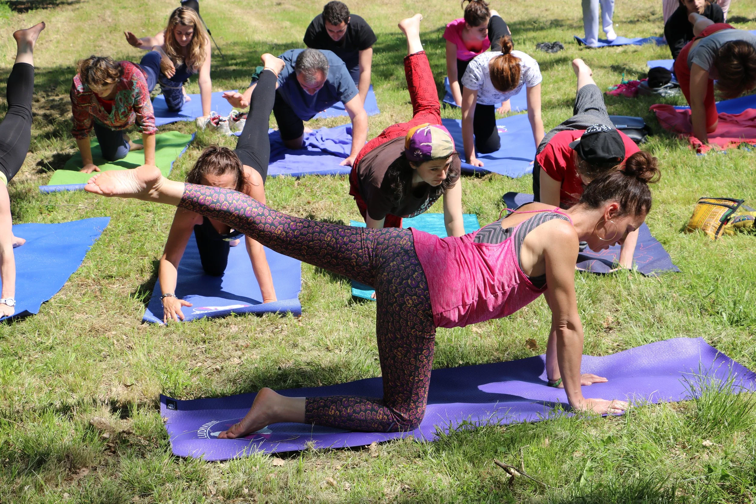 Un groupe de personnes pratiquant le yoga en extérieur sur des tapis colorés dans un endroit verdoyant.