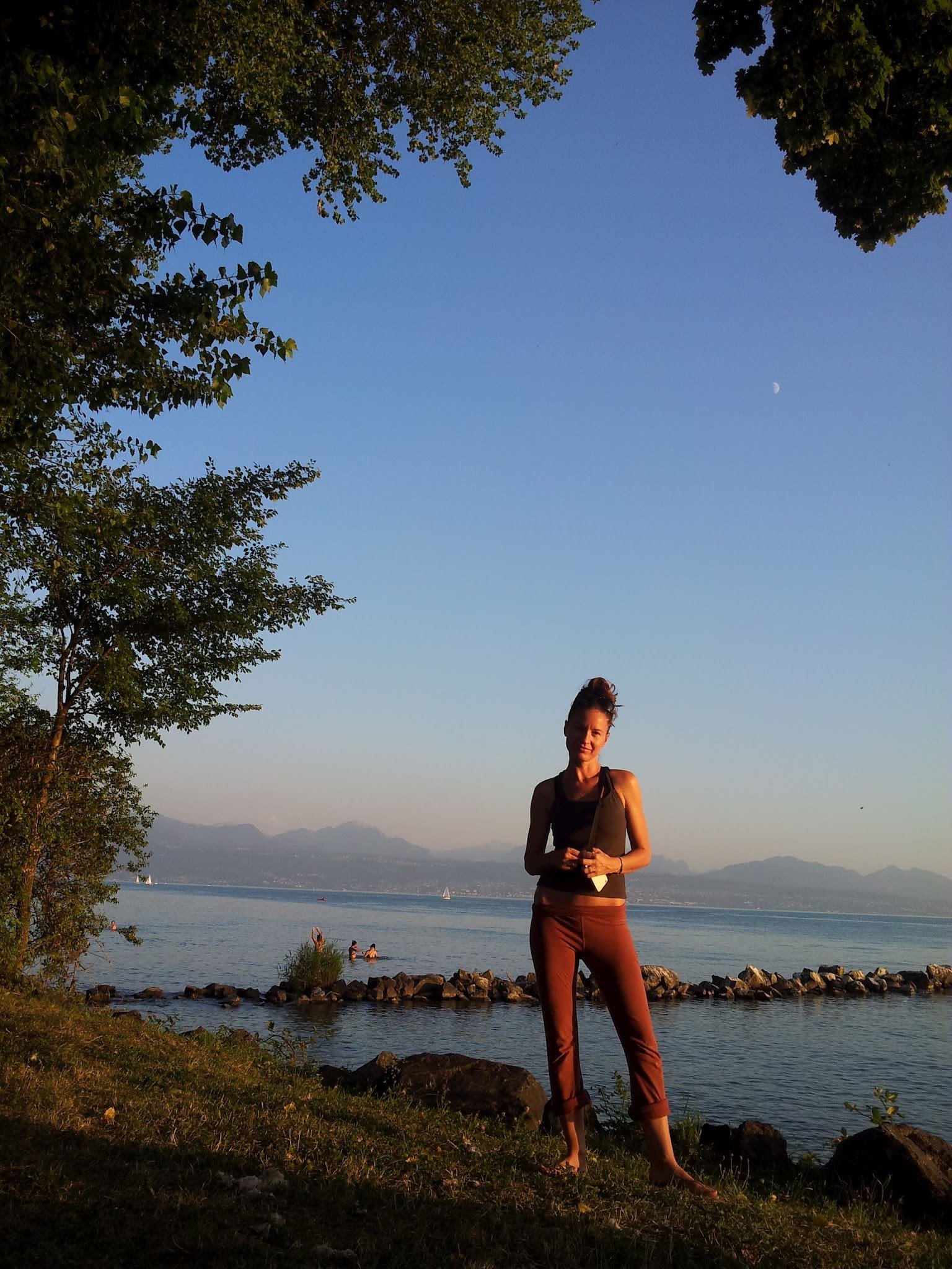 Une femme debout près d'un lac, avec des montagnes au loin, sous un ciel clair avec la lune visible, entourée d'arbres, au coucher du soleil.