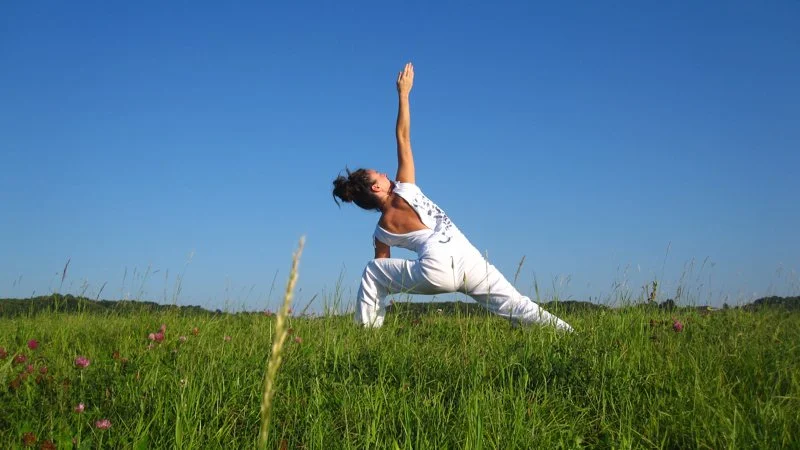Femme pratiquant le yoga en plein air dans un champ, sous un ciel bleu clair.