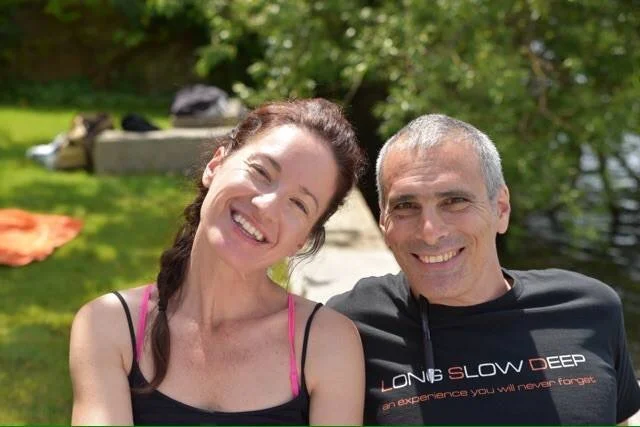 Un couple souriant assis dans un cadre extérieur verdoyant, probablement près d'une rivière ou d'un lac, en train de profiter du soleil.