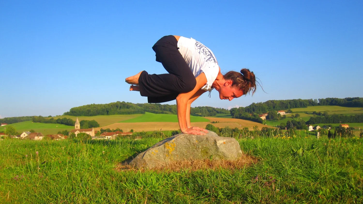 Une femme en position de yoga en équilibre sur ses mains sur un rocher dans un champ vert, avec un paysage de collines et un village au fond.