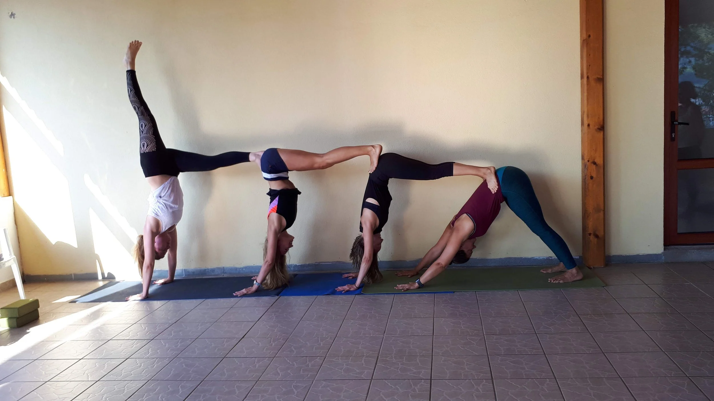 Quatre personnes en position de yoga inversée et de pont formant une chaîne sur des tapis de yoga dans une pièce avec un mur jaune et une porte en bois.