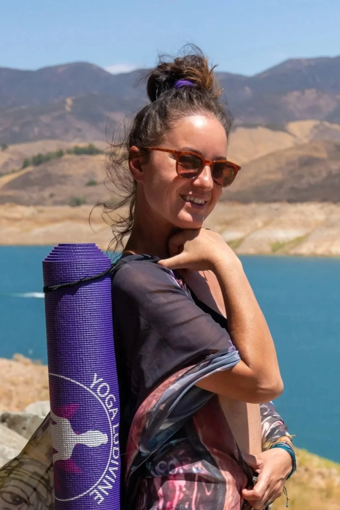 Une femme souriante portant des lunettes de soleil, un sac de yoga violet sur le dos, en plein air avec un lac et des montagnes en arrière-plan en Californie.