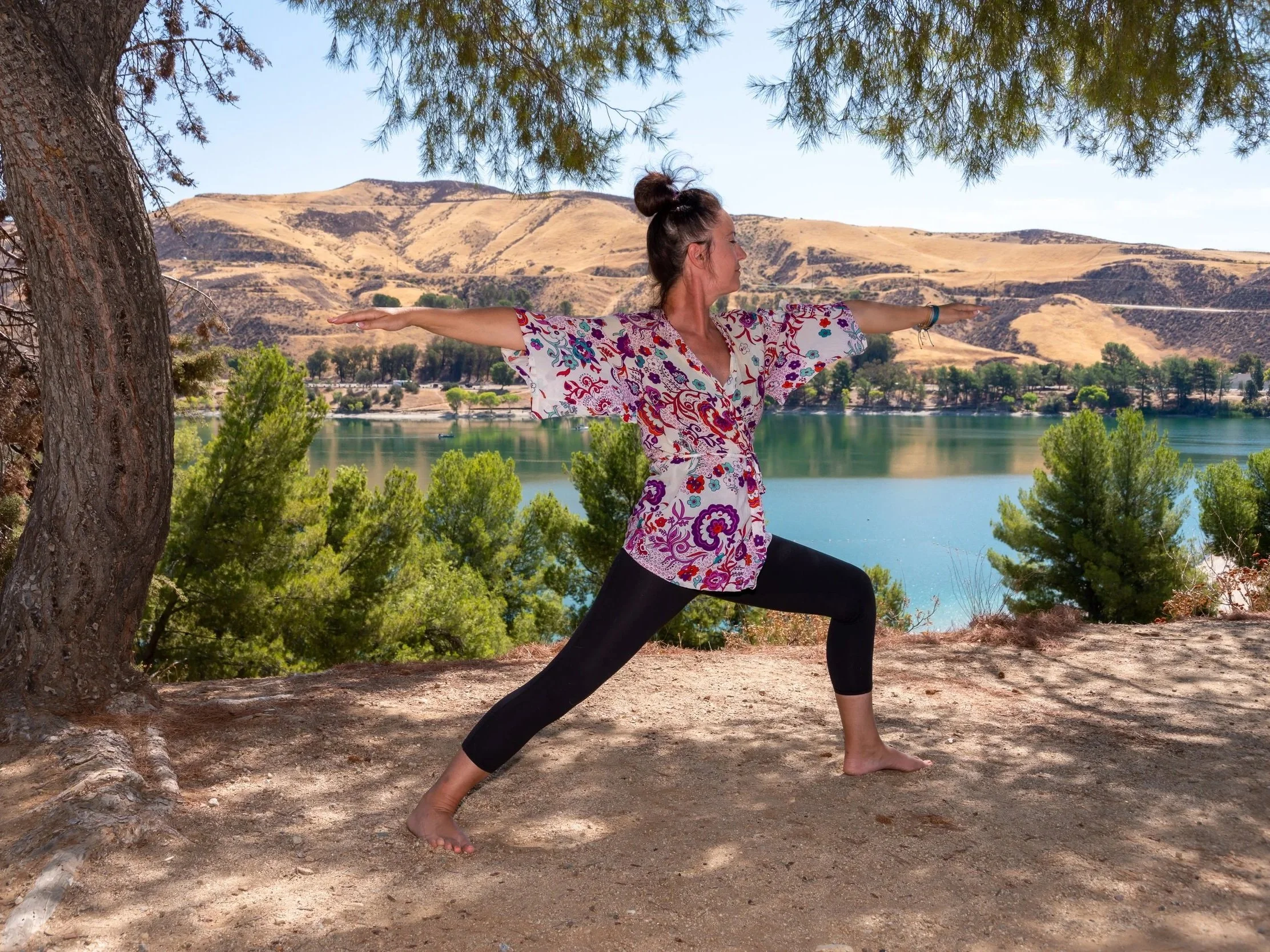 Femme faisant du yoga dans un parc près d'un lac avec des montagnes en arrière-plan, portant un kimono fleuri et leggings noirs, sous un arbre.