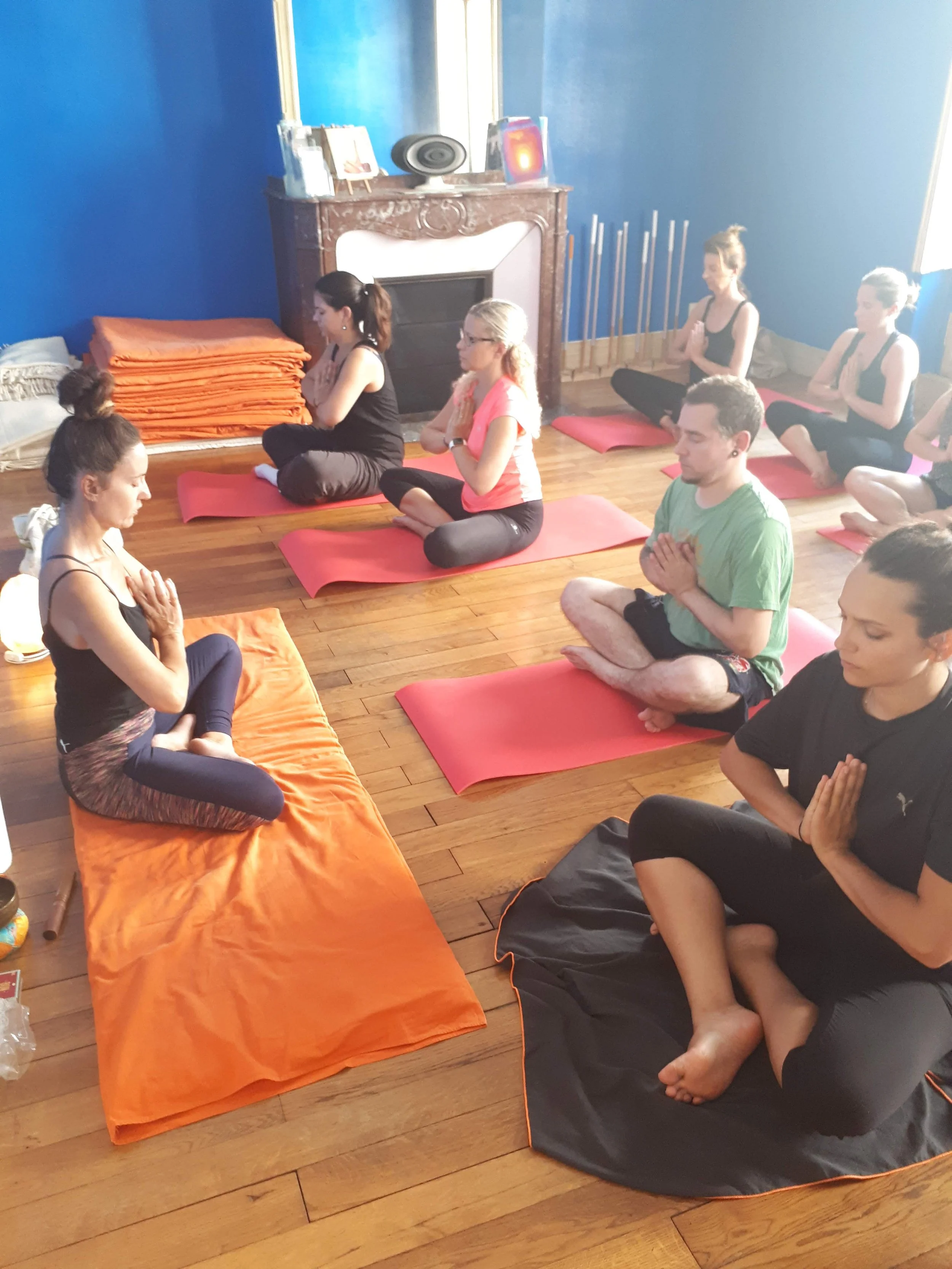 groupe de personnes assises sur des tapis de yoga dans une salle de yoga, pratiquant la méditation ou la relaxation, avec une cheminée en arrière-plan et des fenêtres laissant entrer de la lumière naturelle.