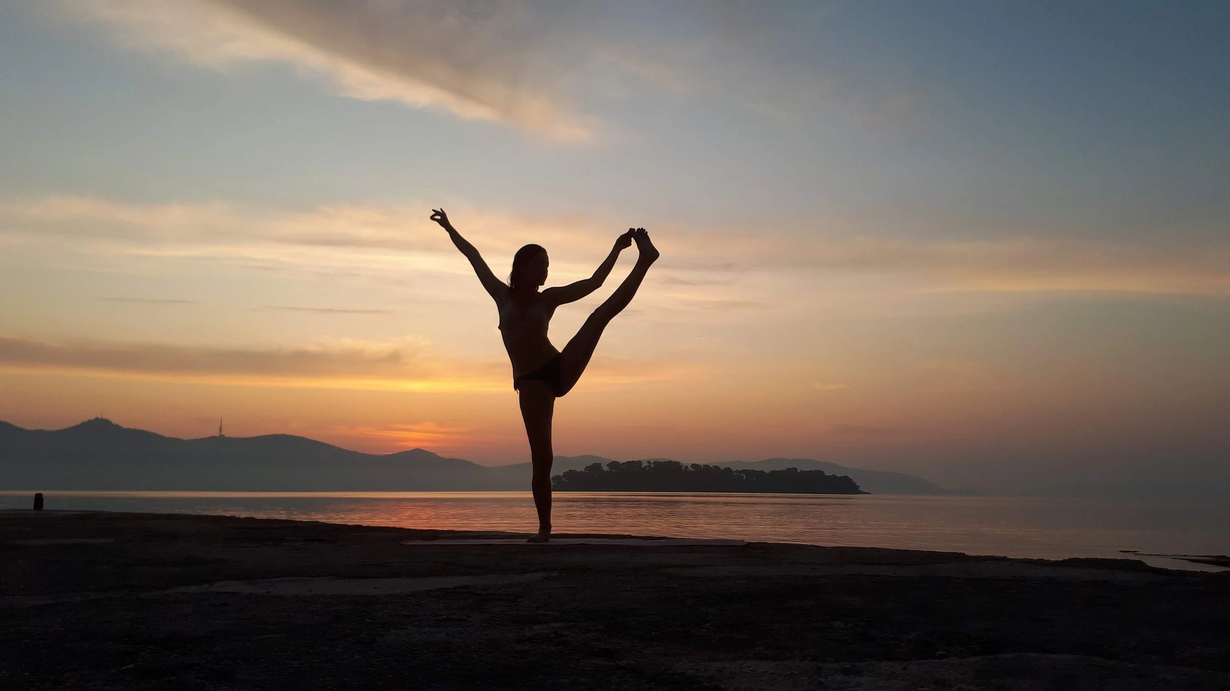 Silhouette d'une femme faisant une pose de yoga sur une plage au coucher du soleil, avec des montagnes à l'horizon.
