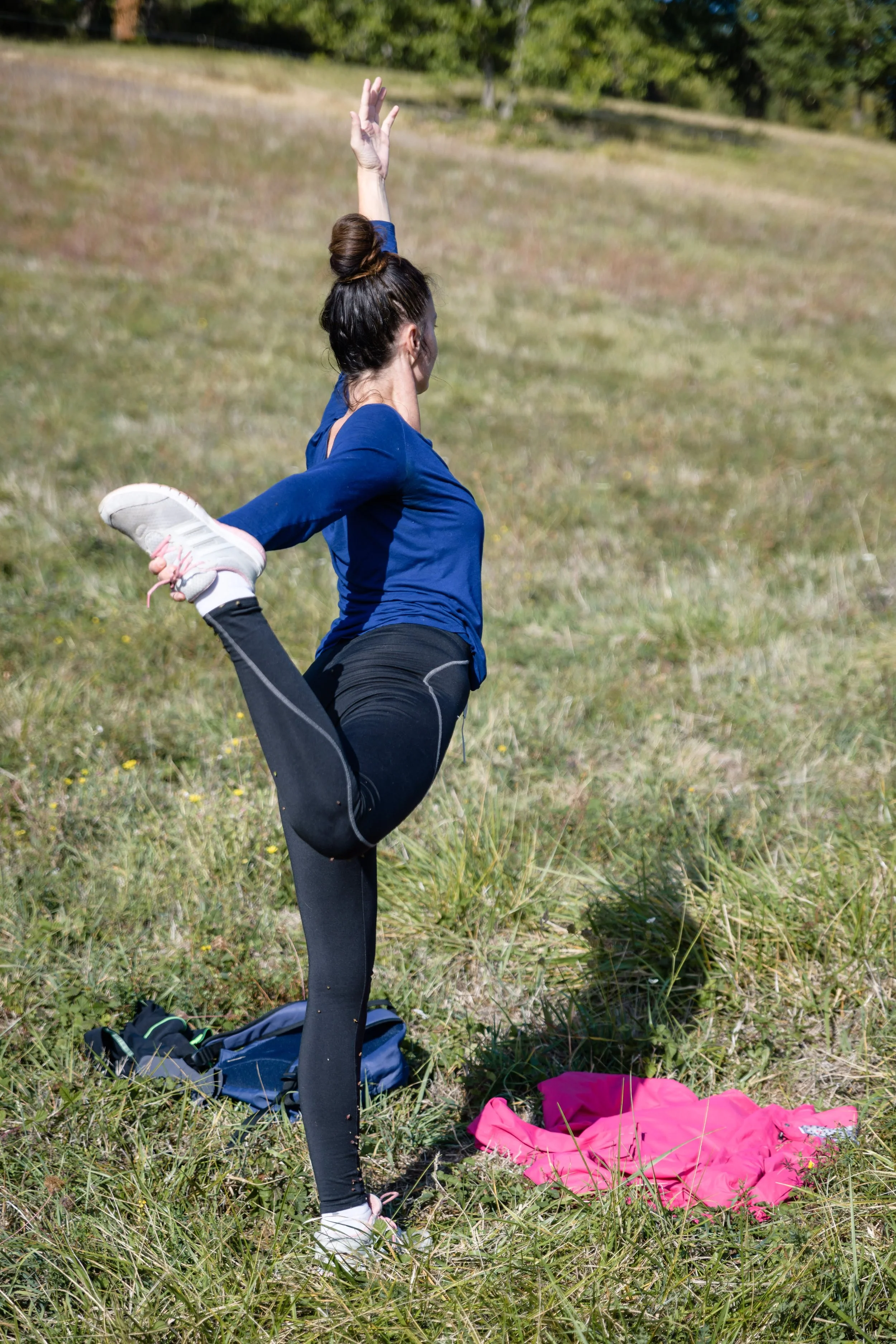 Une femme en tenue de sport pratique une posture de yoga dans un champ herbeux, avec des sacs à dos et un vêtement rose sur le sol à proximité.