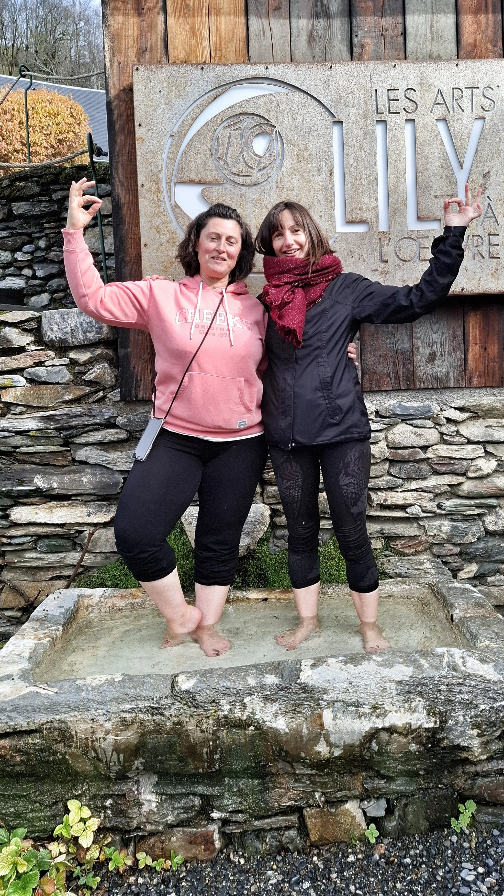 Deux femmes souriantes, pieds dans l'eau, posent sous un panneau en pierre marquant l'entrée à Lily à l'Océan, avec un mur en pierre en arrière-plan.