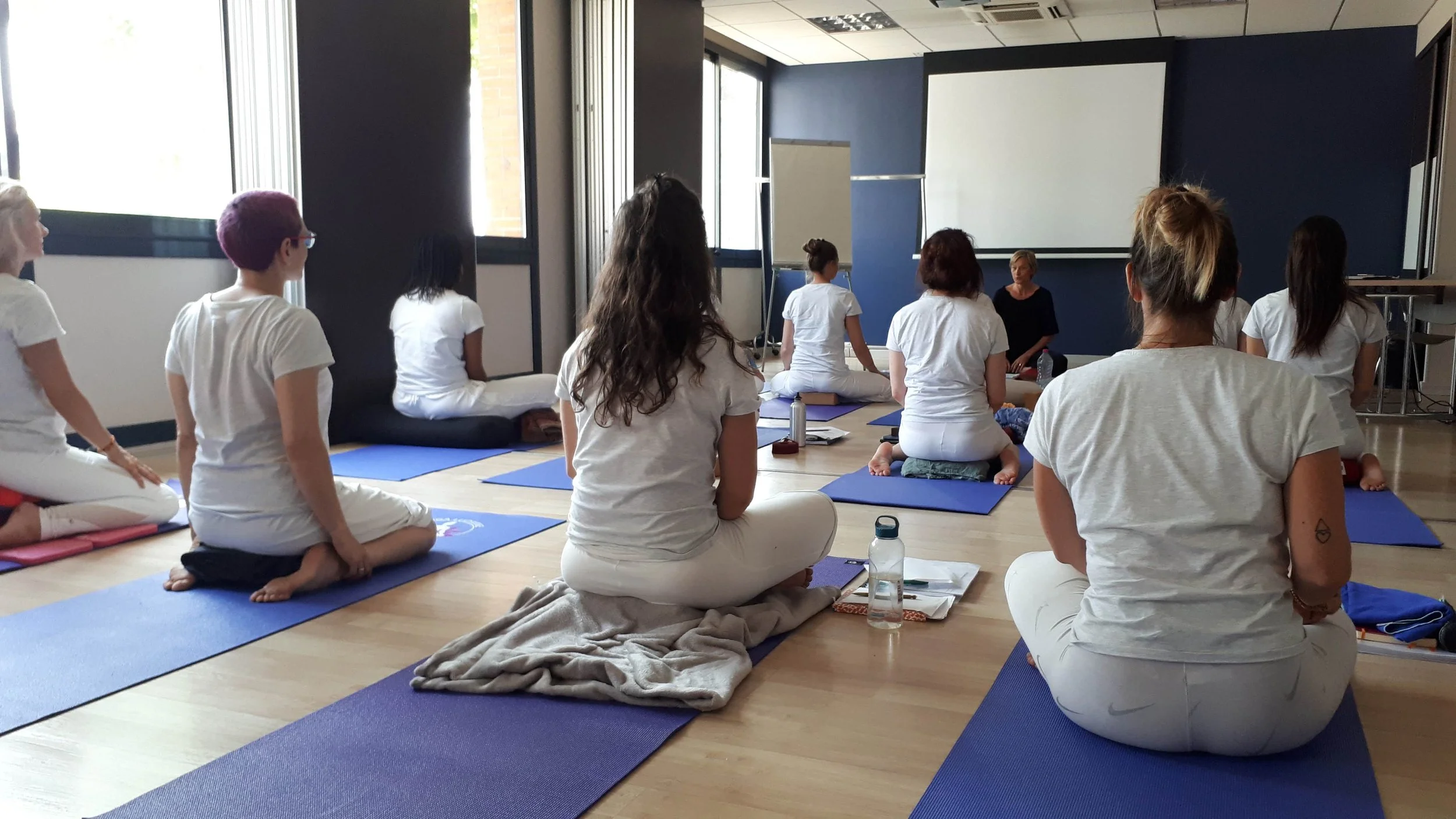 Groupe de femmes en vêtements de yoga assises en position de méditation sur des tapis bleus dans une salle lumineuse, avec une femme en position de instructrice ou guide devant elles.