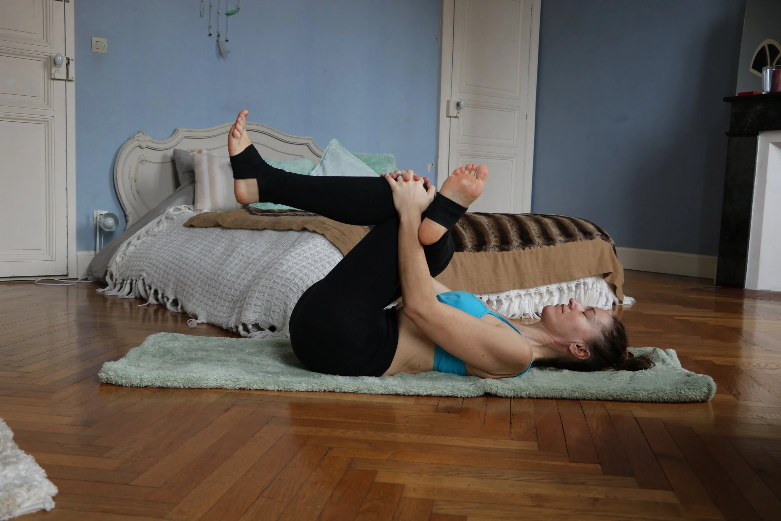 Femme faisant un exercice de yoga allongée sur un tapis, genoux pliés, mains sur les jambes, dans une chambre