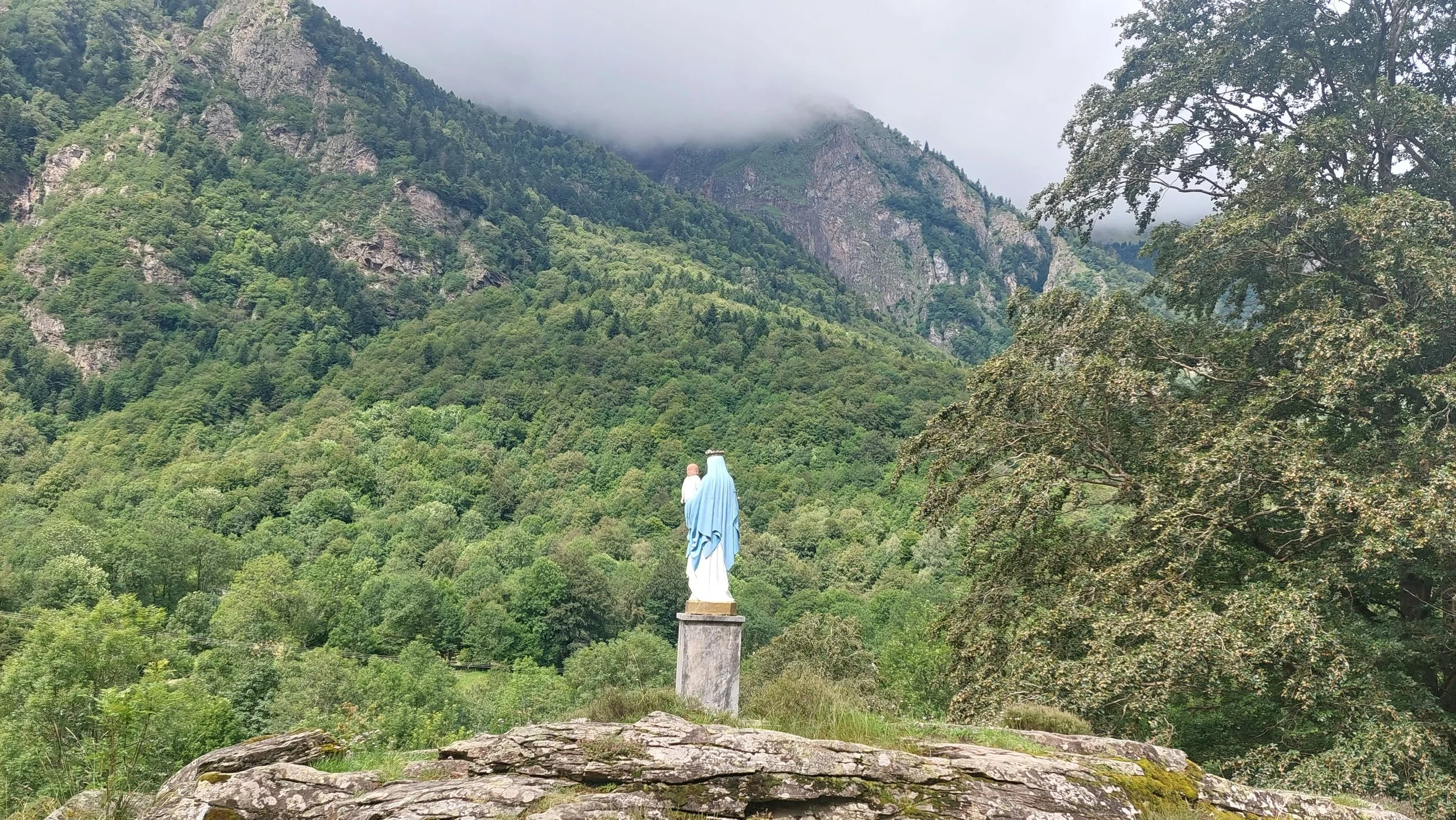 Statue religieuse de la Vierge Marie, vêtue de bleu et blanc, située sur un rocher face à une forêt dense avec des montagnes en arrière-plan, sous un ciel nuageux.