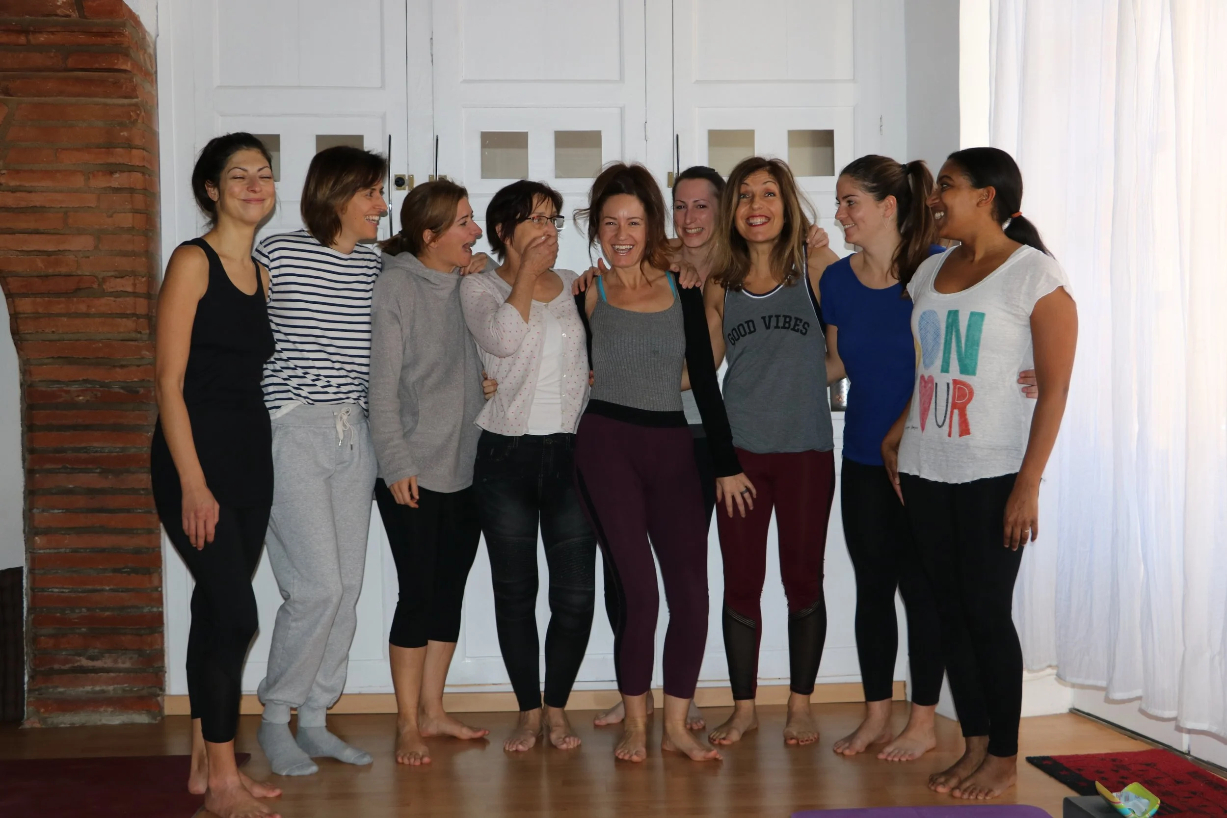 groupe de femmes souriantes posant ensemble dans une pièce avec sol en bois, mur blanc et cheminée en briques, certaines en vêtements de sport, à l'air détendu et heureux.