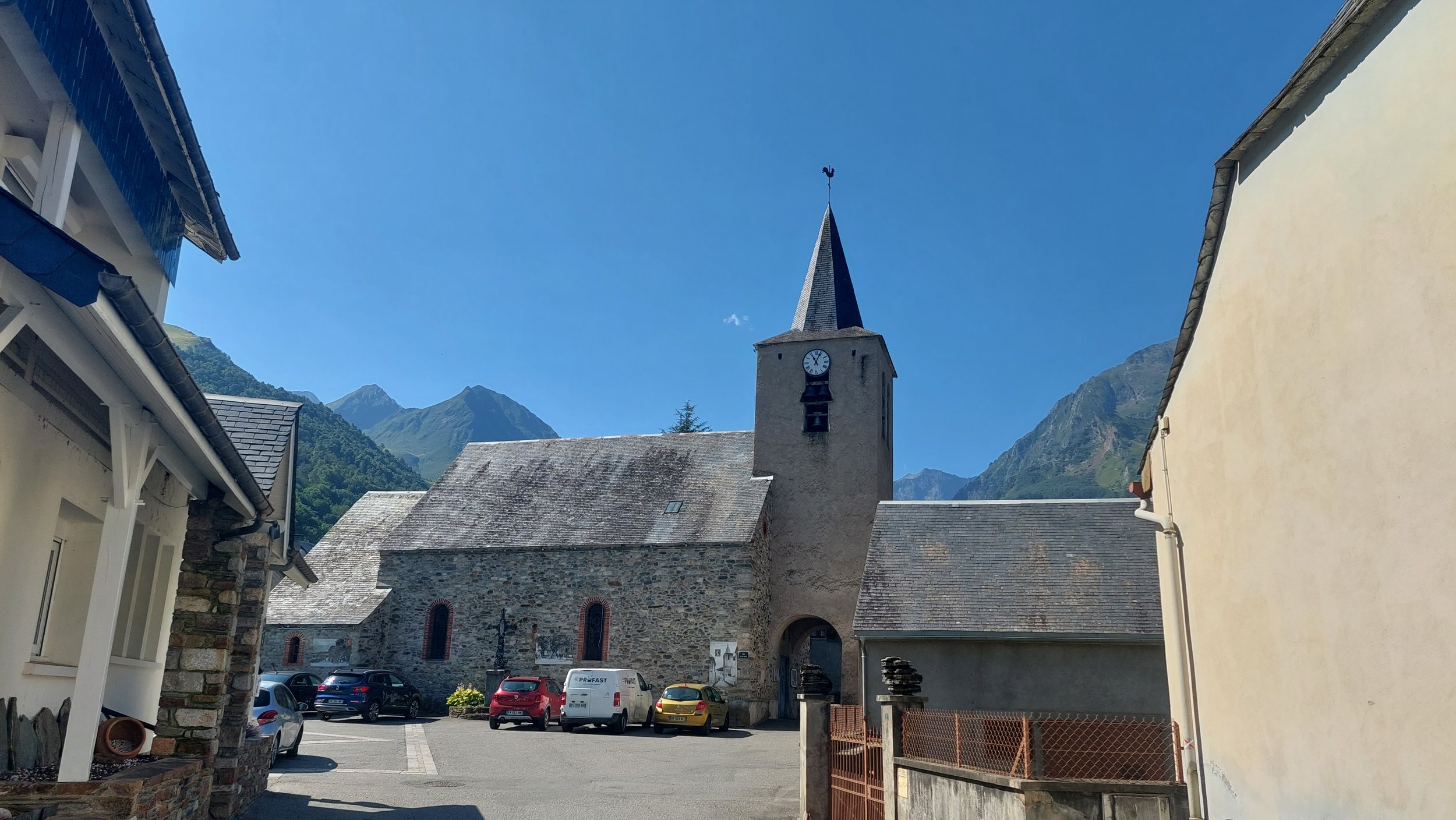 Une église en pierre avec un clocher pointu, entourée de montagnes, face à un parking avec plusieurs voitures, sous un ciel bleu clair.