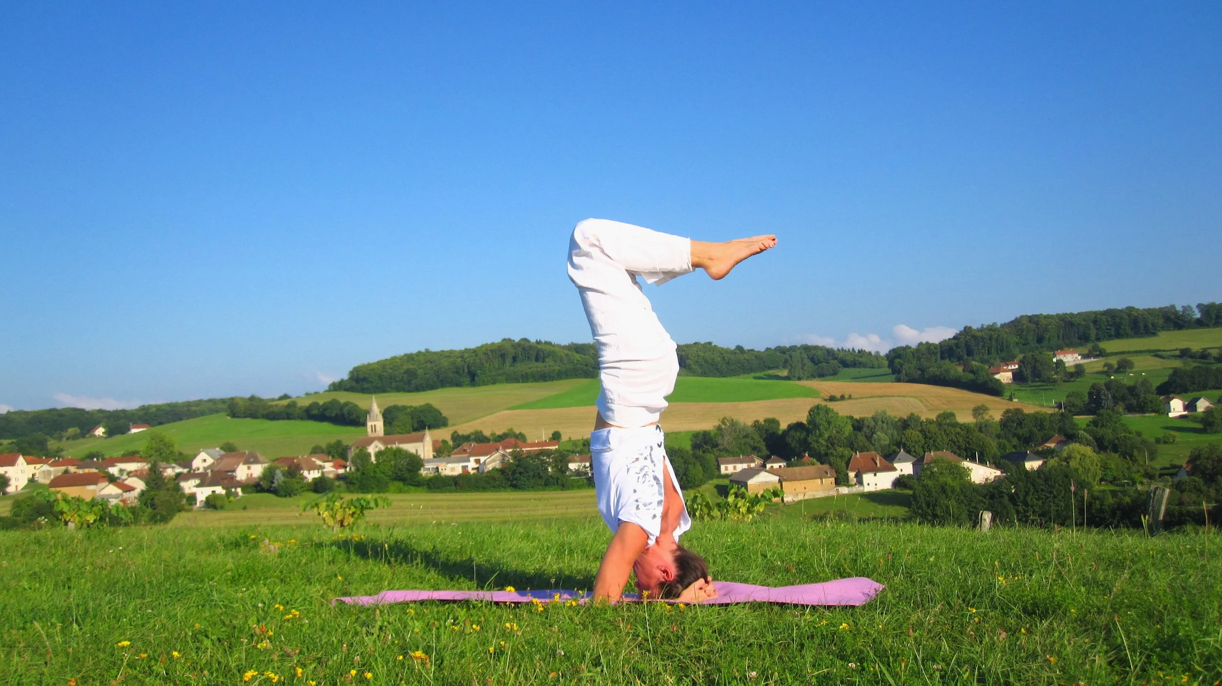 Une personne pratiquant le yoga en position sur la tête dans un champ vert avec un village et des collines en arrière-plan, sous un ciel bleu clair.