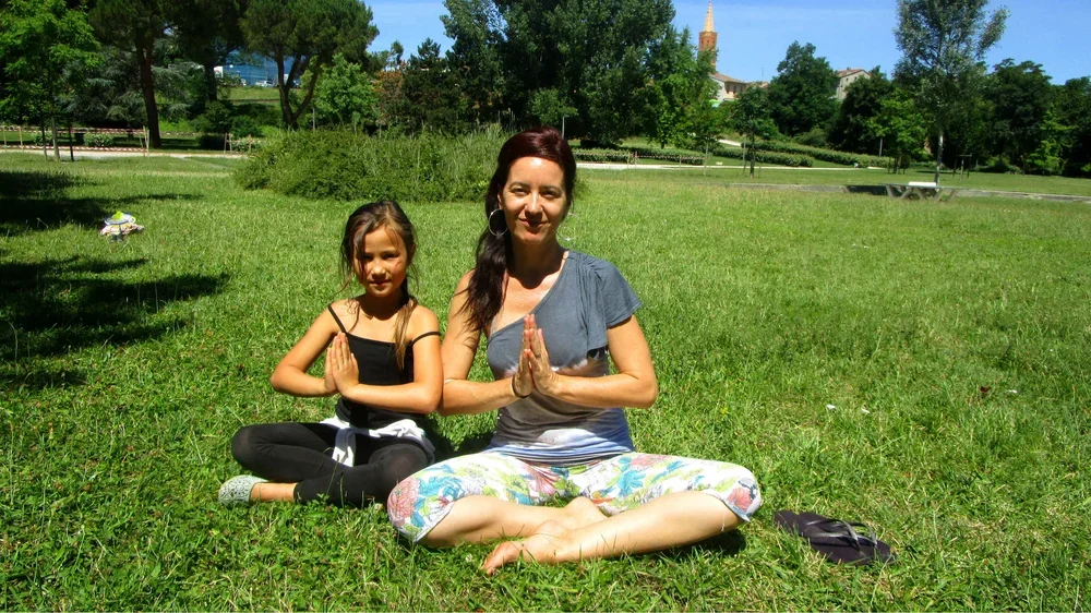 Une femme et une jeune fille pratiquant la méditation ou le yoga en position de lotus dans un parc ensoleillé, avec des arbres verts et un ciel bleu.