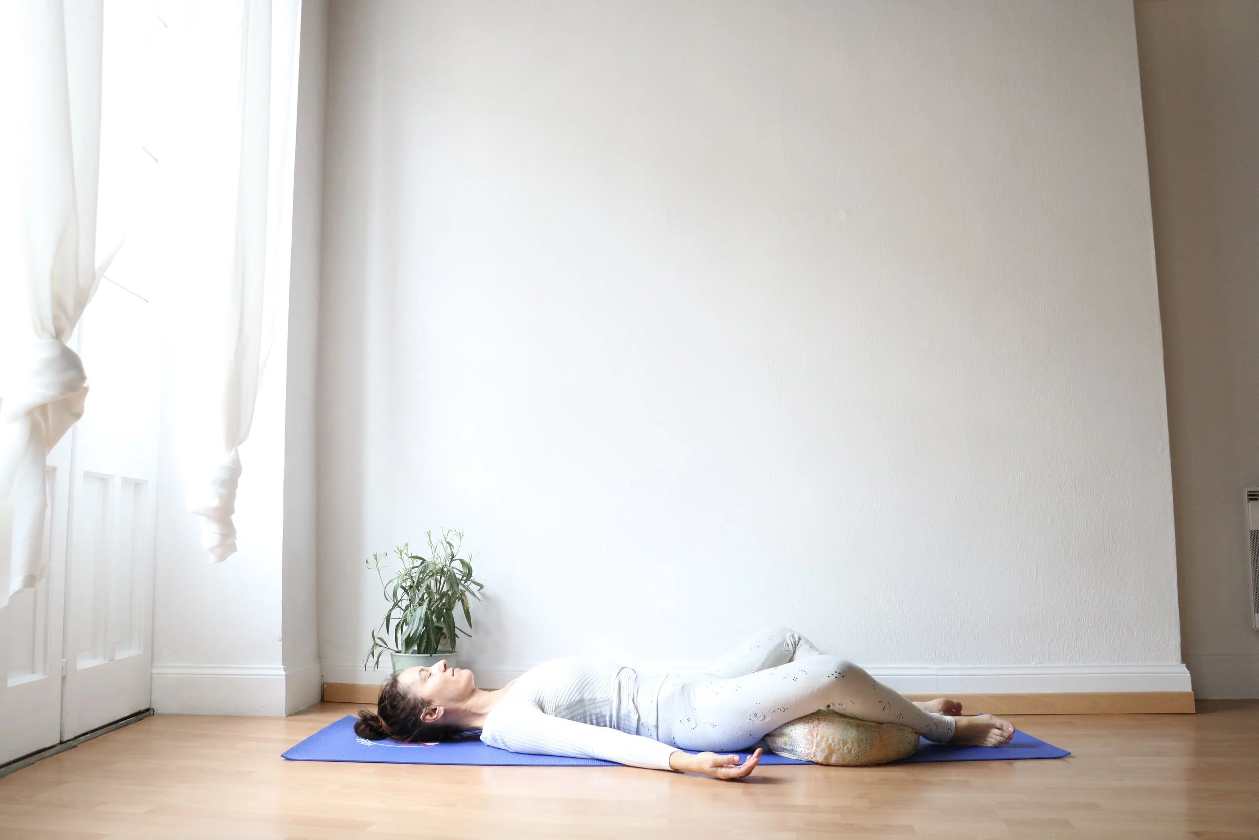 Femme faisant une posture de relaxation ou de yoga allongée sur un tapis bleu dans une pièce lumineuse avec une plante verte à côté.