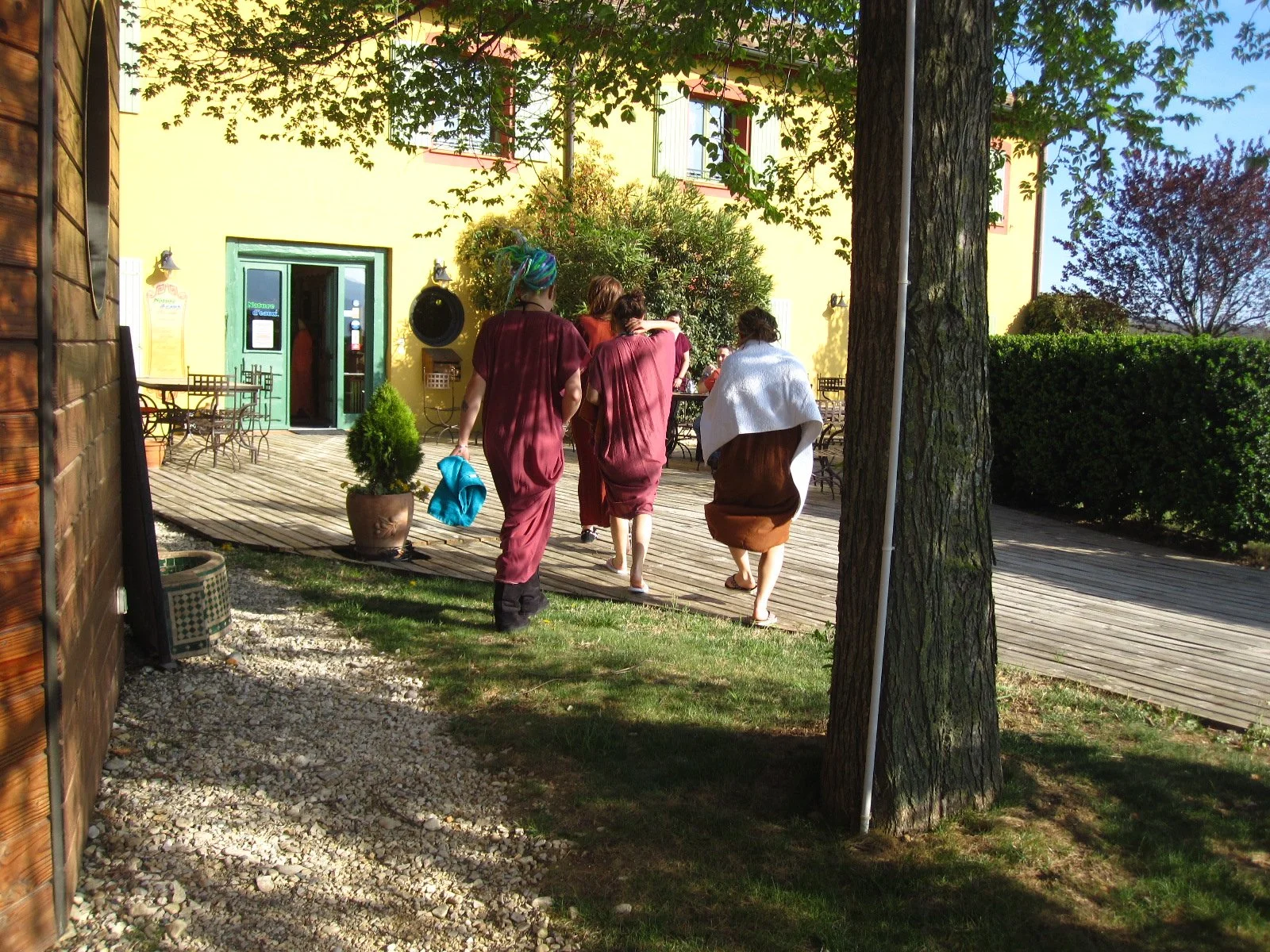 Groupe de personnes marchant vers un bâtiment jaune avec une porte verte, entouré d'arbres et de buissons, en plein jour.