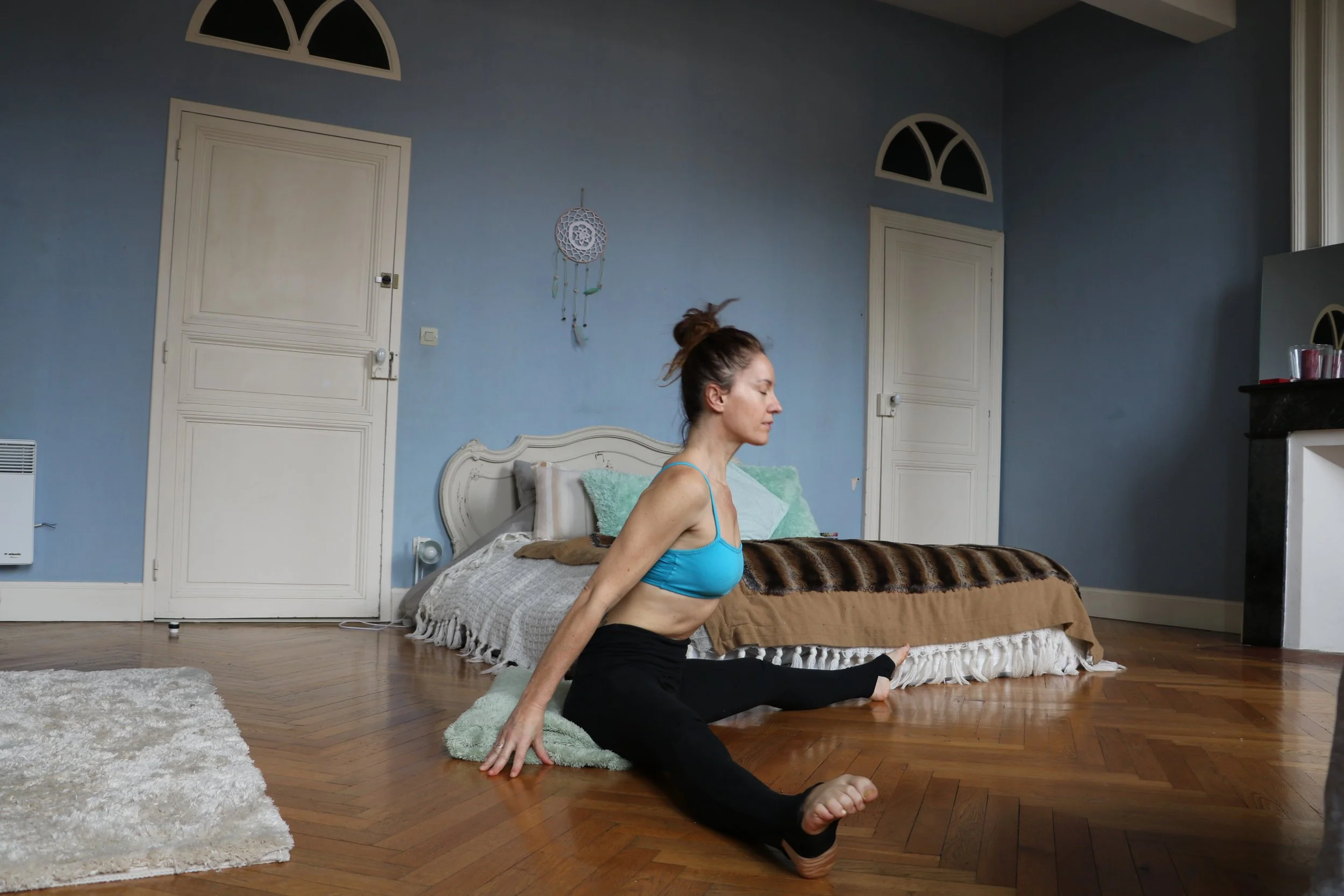 Une femme en vêtements de sport pratique le yoga ou des étirements dans une chambre avec un parquet en bois et un lit en arrière-plan.