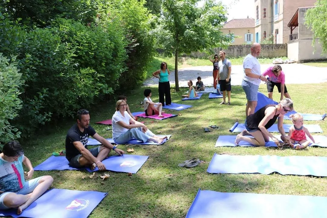 Groupe de personnes assises sur des tapis de yoga dans un parc, quelques personnes se tiennent debout, certains enfants sont assis, en train de participer à une séance de yoga en plein air