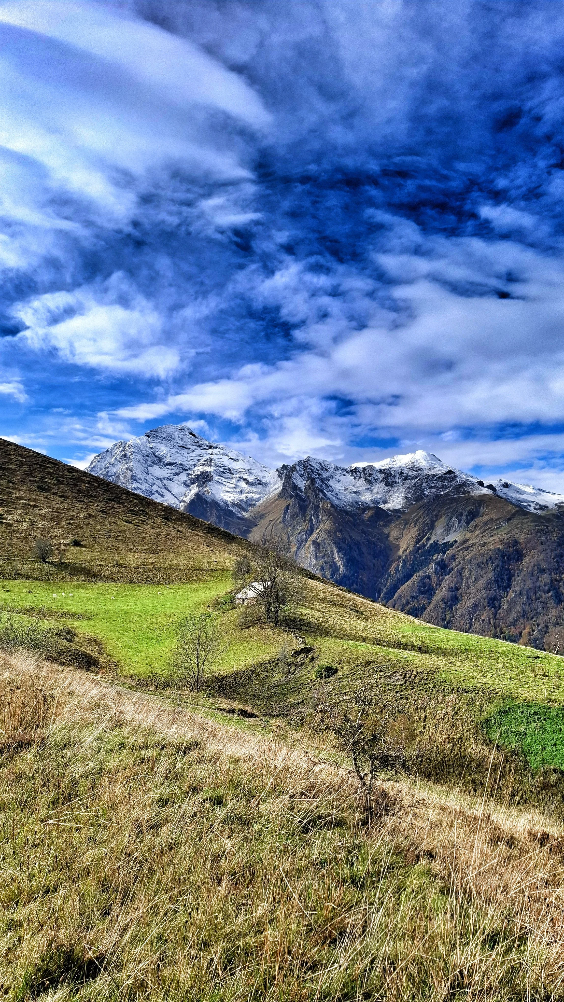Paysage de montagnes enneigées sous un ciel bleu avec des nuages, près d'une vallée verte avec quelques arbres et une petite ferme.