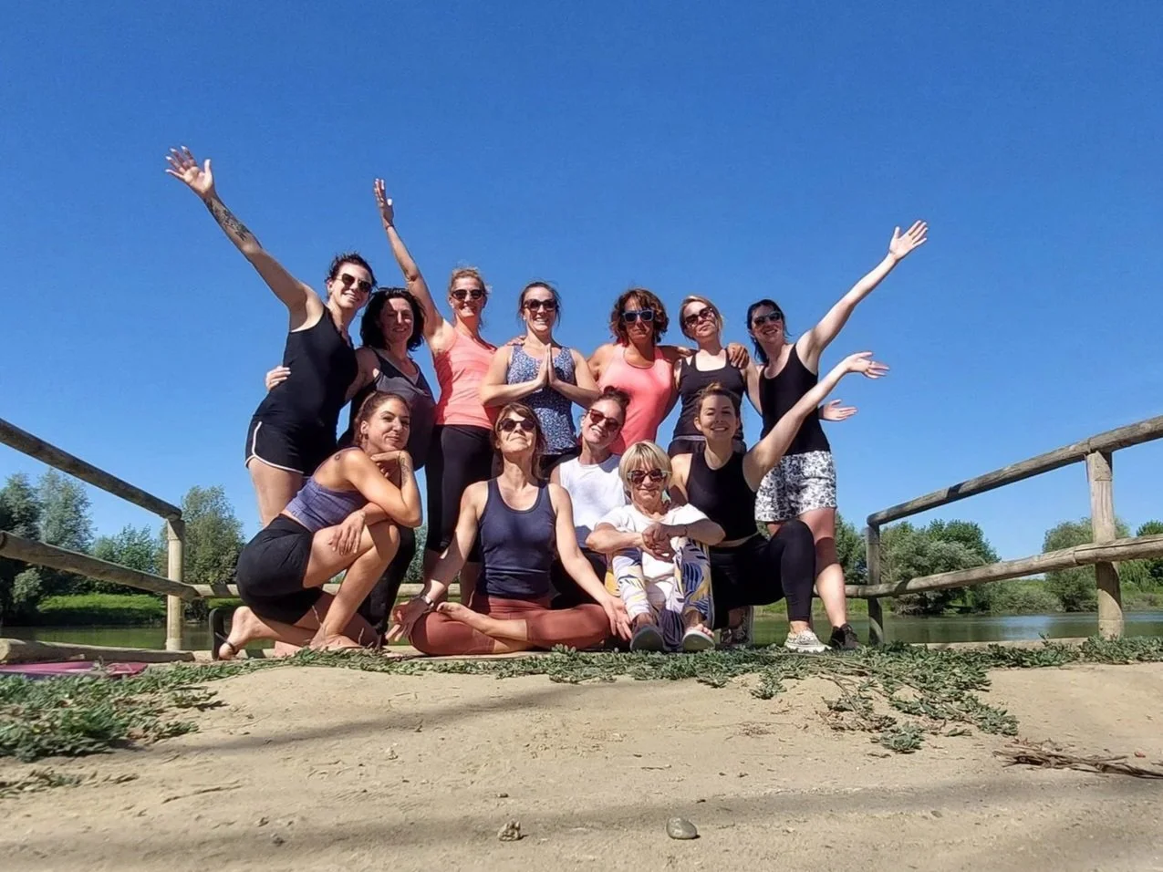 Groupe de femmes posant ensemble sur un pont en bois en plein air, sous un ciel bleu, face à une rivière et des arbres, lors d'une sortie ou d'une activité de groupe par beau temps. Durant une formation de yoga.