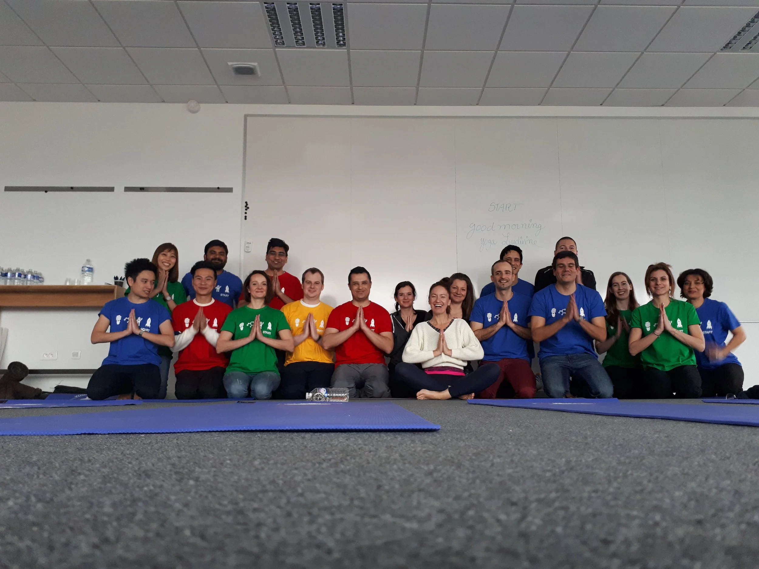 Groupe de personnes assises sur le sol en tapis de yoga, faisant la position de prière avec les mains, dans une salle blanche avec un tableau derrière et des t-shirts colorés (bleu, rouge, vert, jaune)