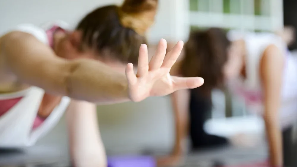 Trois personnes pratiquent la pose de la planche pendant une séance de yoga ou de méditation en intérieur, avec une personne en premier plan qui étend sa main.