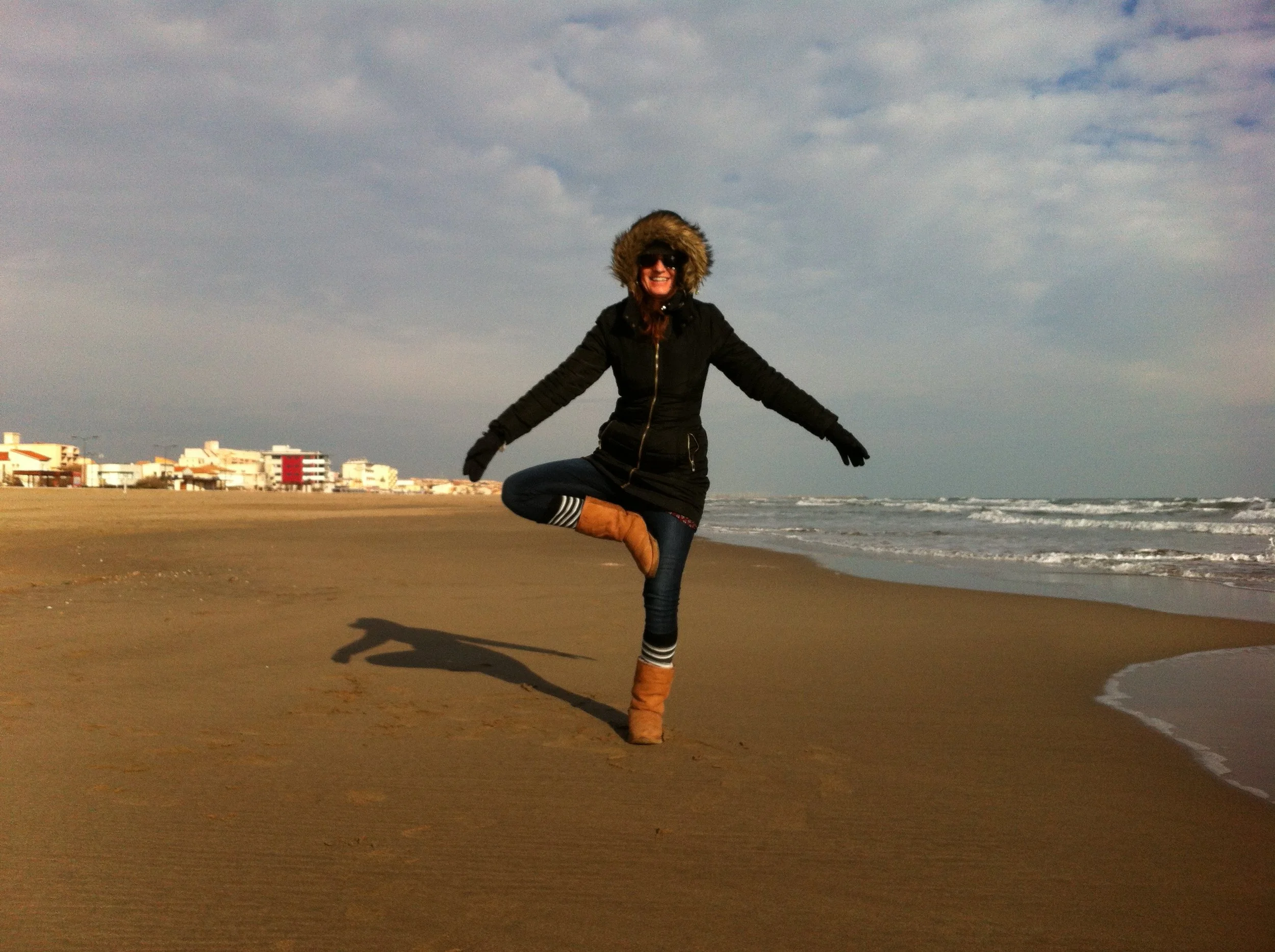 Ludivine souriante en manteau noir et bottes en daim, faisant du yoga position de l'arbre sur la plage à Arcachon au coucher du soleil, avec des bâtiments en arrière-plan et des vagues dans l'océan.