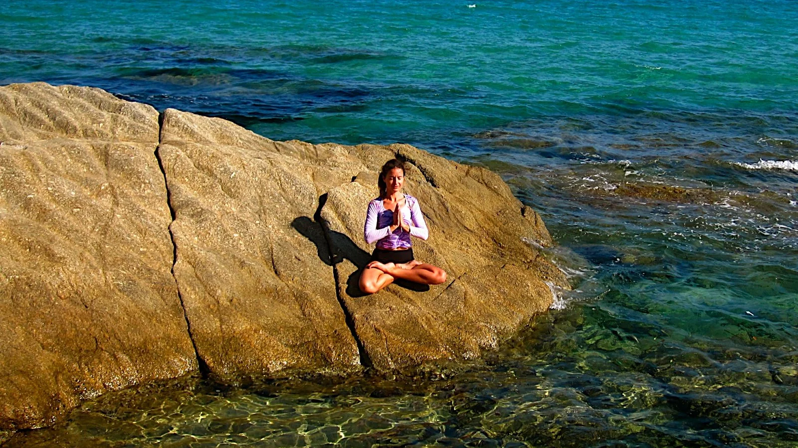 Une femme assise en position de méditation sur une roche au bord de la mer, en tailleur, avec les mains en prière. La mer est calme avec des eaux bleues et vertes.