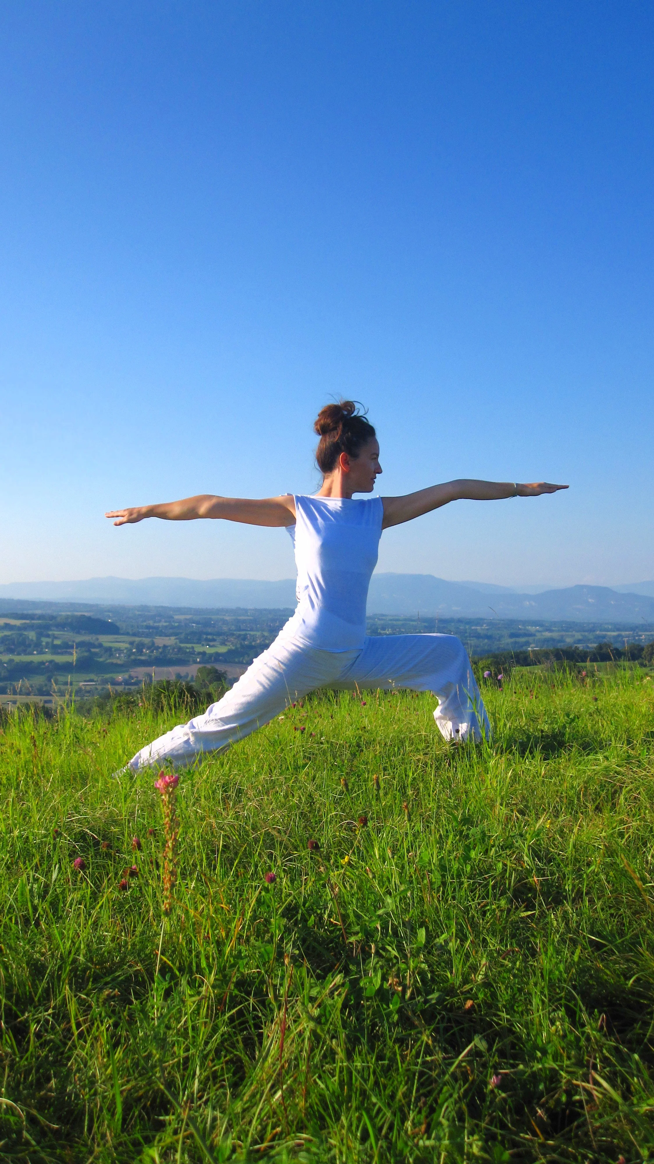 Femme faisant du yoga en posture de guerrier dans un champ vert avec une vue sur la campagne et des montagnes au loin, ciel bleu clair.