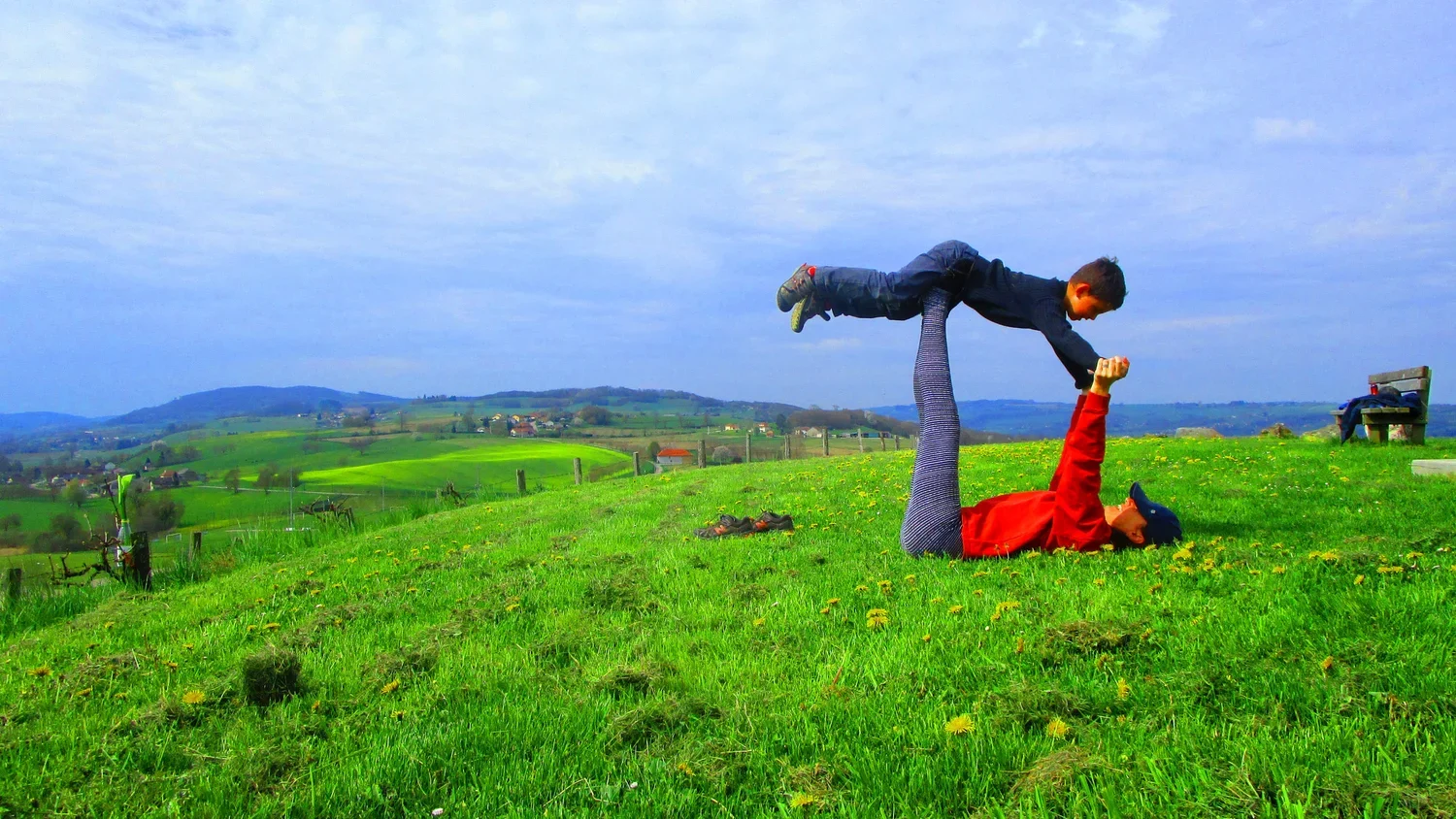 Yoga pour les enfants, professeur et élève pratiquant une acrobatie dans un champ vert avec un paysage de collines verdoyantes en arrière-plan. L'une est allongée sur le dos, tenant l'enfant en position de yoga : avion
