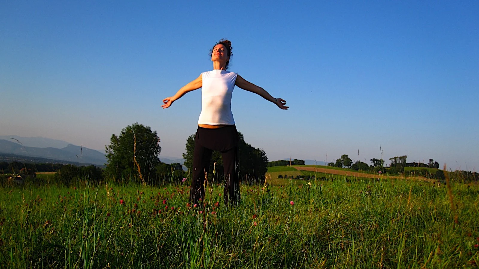 Femme debout dans un champ vert avec les bras étendus, sous un ciel clair, paysage de campagne avec des arbres et des montagnes en arrière-plan.