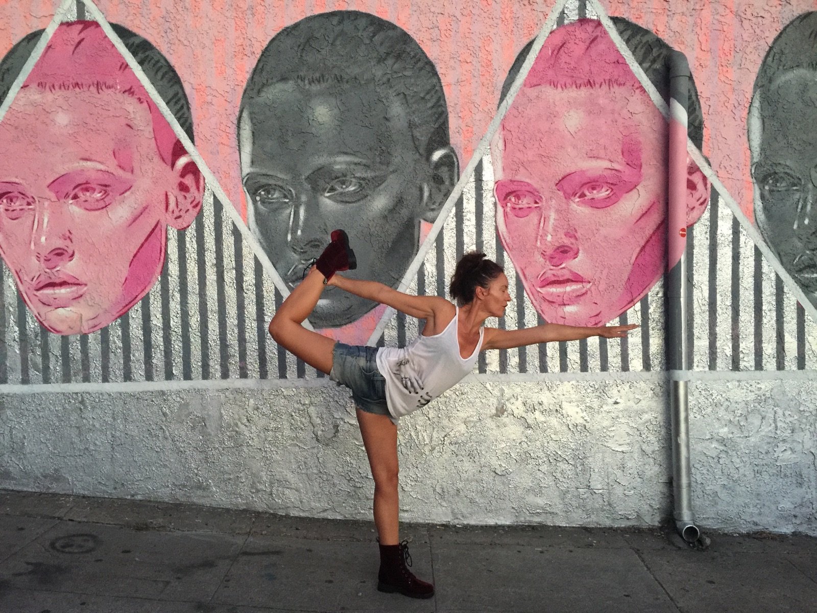 Une femme fait une pose de yoga en équilibre sur une jambe devant un mur orné d'un graffiti représentant plusieurs visages en nuances de gris et de rose.