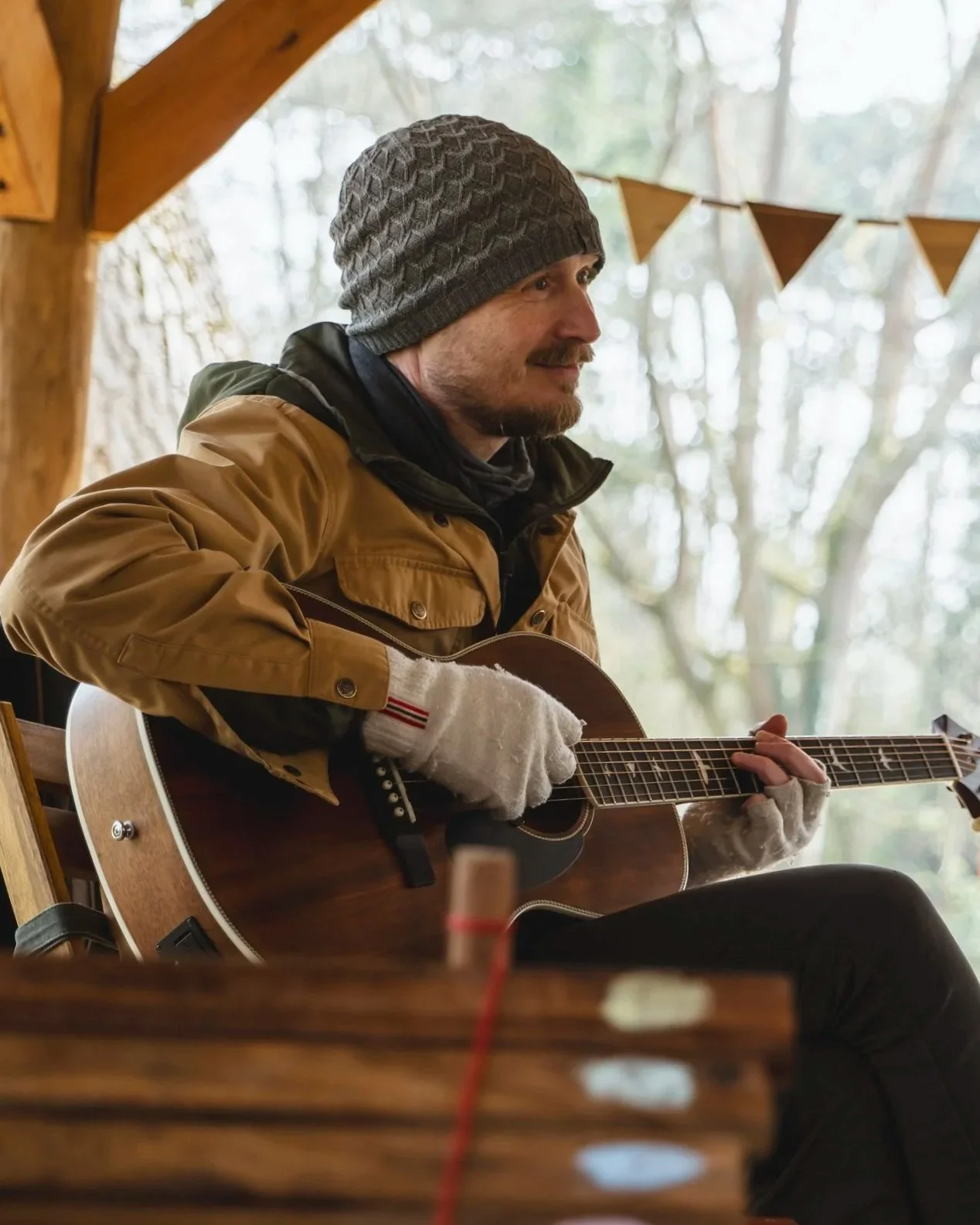 A man wearing a grey knit beanie, brown jacket, and white gloves, is sitting and playing an acoustic guitar indoors near a wooden structure with window view of trees.