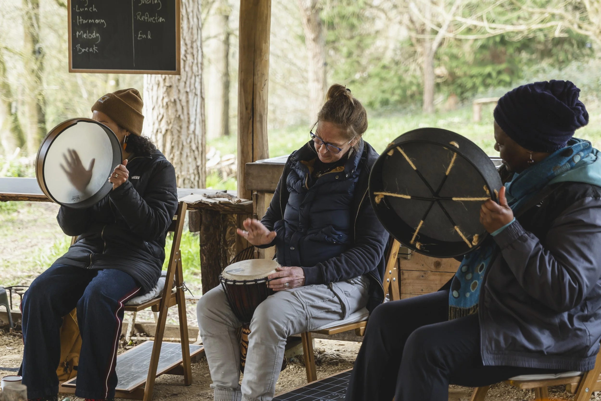 Three women sitting outdoors, playing hand drums in a wooded area with a blackboard in the background.