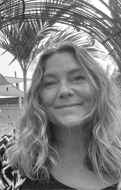 A woman with wavy hair smiling outdoors, with palm leaves and a shaded area in the background.