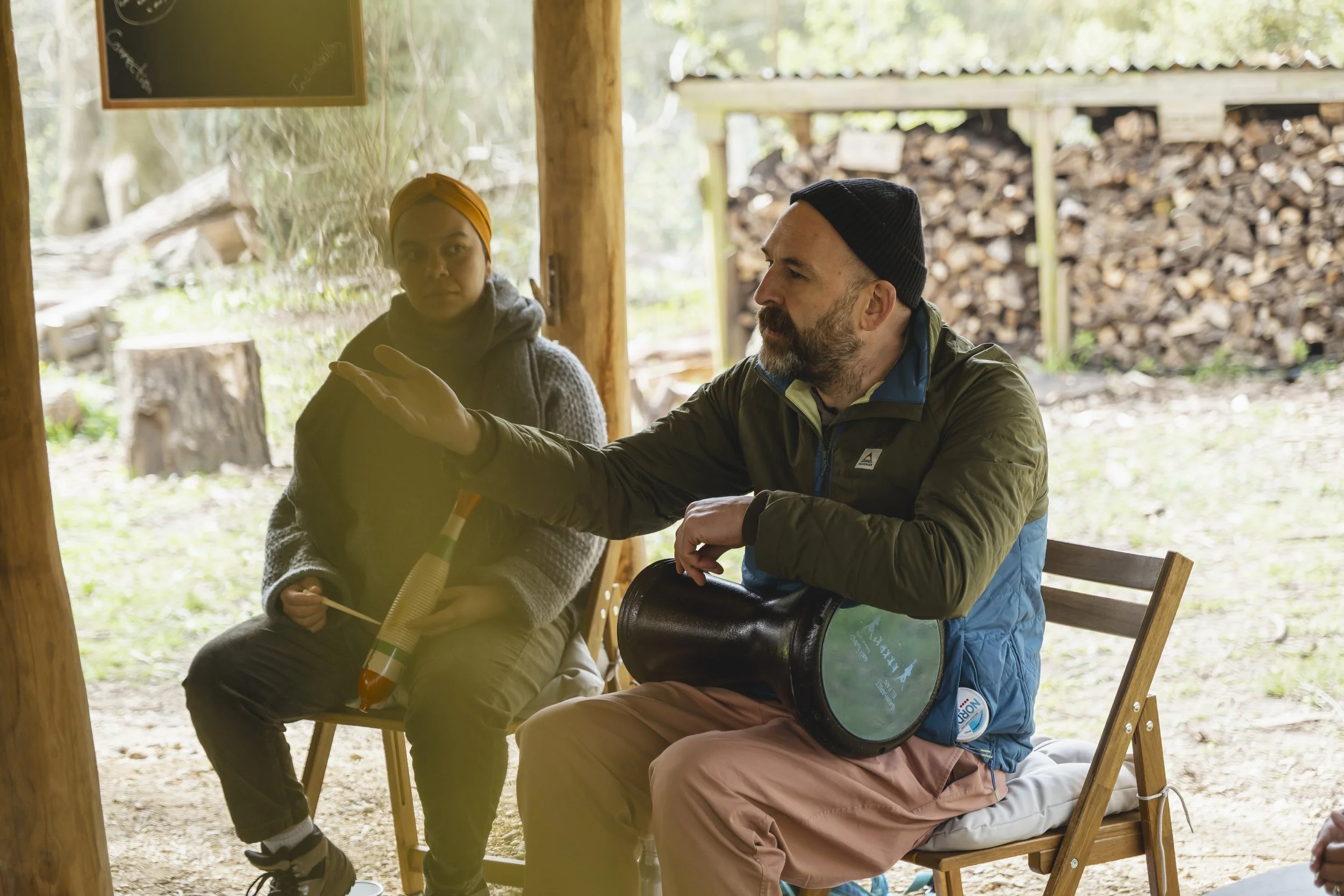 Two people sit outside, engaging in a conversation. The man has a beard, wears a black beanie and a green jacket, and is gesturing with his hand. The woman wears a yellow head wrap and a gray sweater, holding a topped object in her hands. There is a wooden structure and a woodpile in the background.