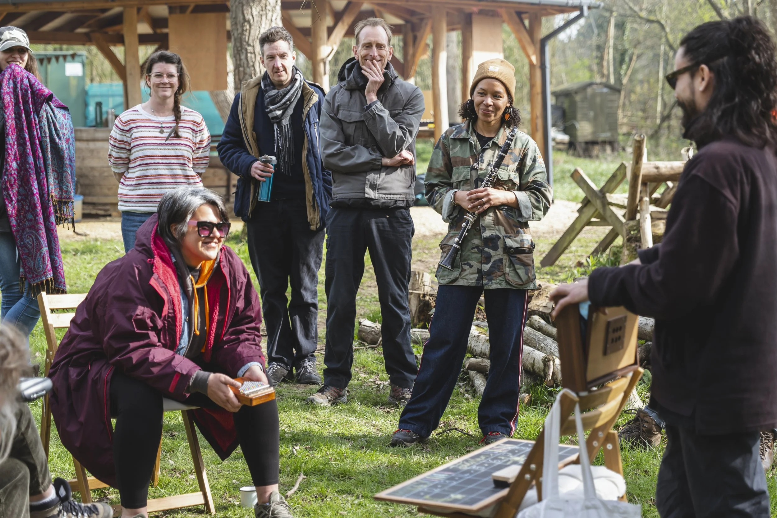 Group of people gathered outdoors in a natural setting, some standing and some sitting, engaged in a conversation or presentation, with trees and wooden structures in the background.